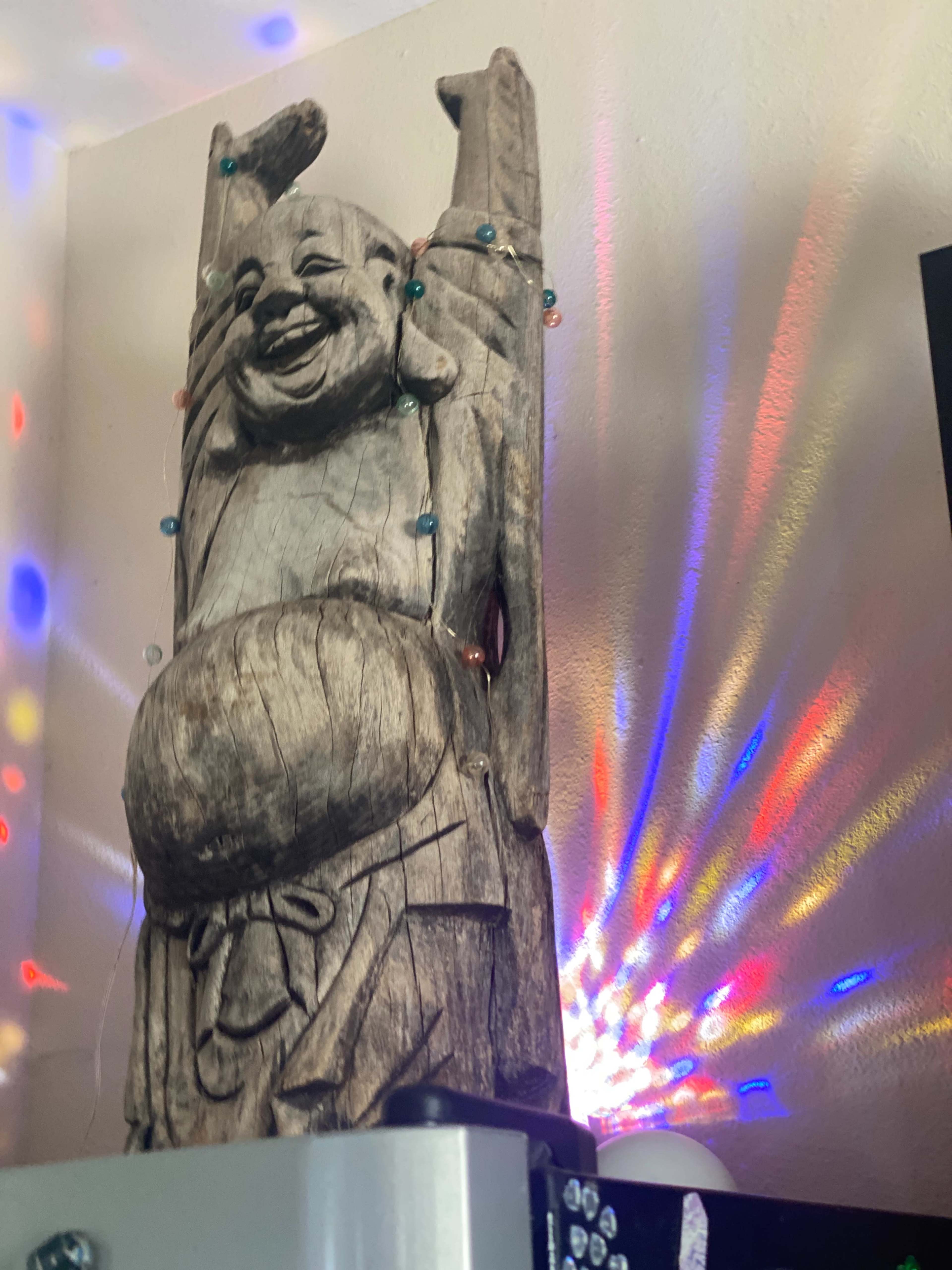A wooden statue of a laughing figure is adorned with colorful lights, standing against a wall that reflects vibrant patterns.