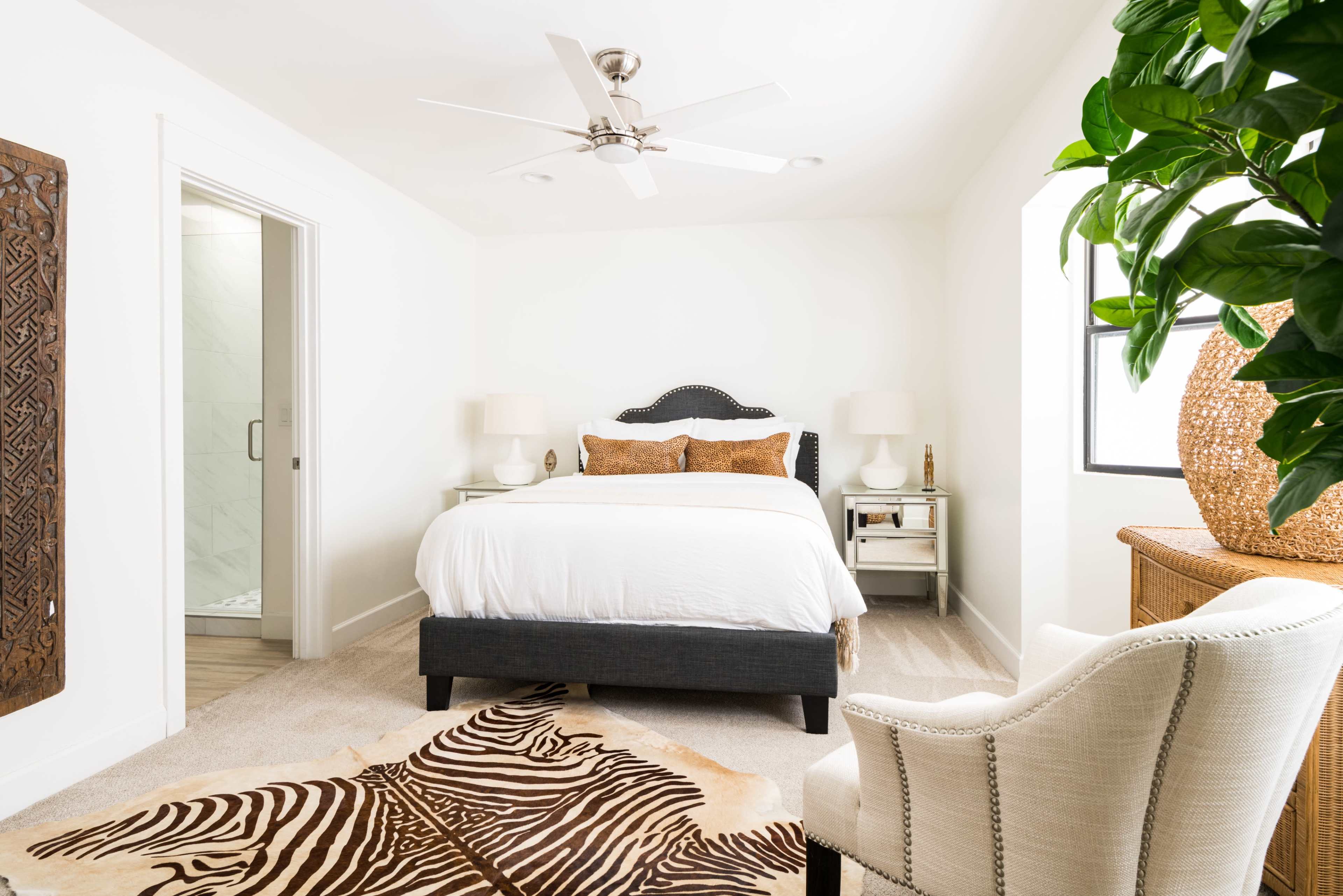 A bright bedroom featuring a white bed, two bedside tables with lamps, a chair, a patterned rug, and a large potted plant.