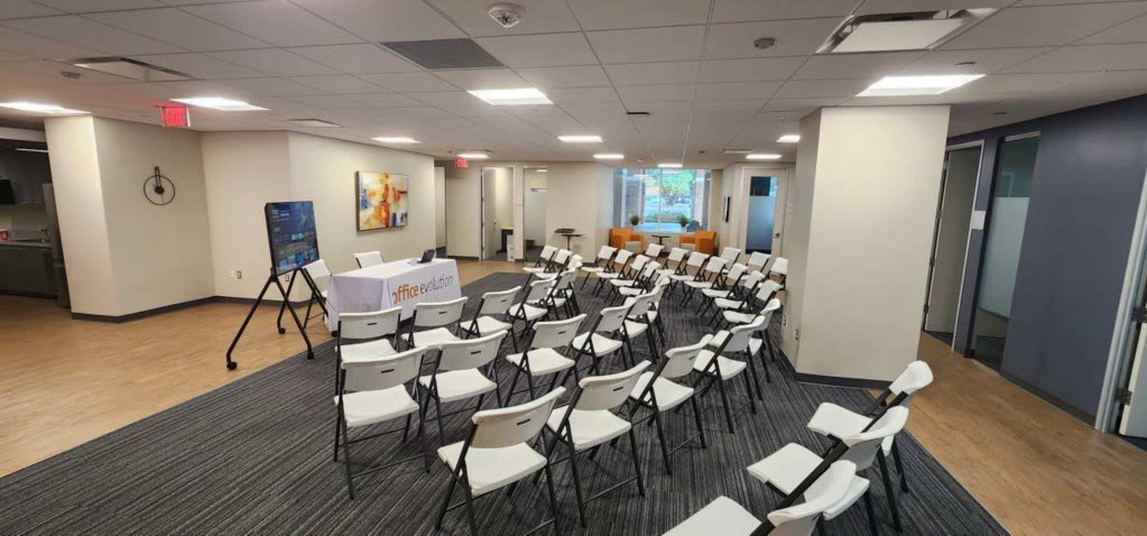 A room is set up with rows of white chairs facing a table adorned with a tablecloth, positioned near large windows with natural light.