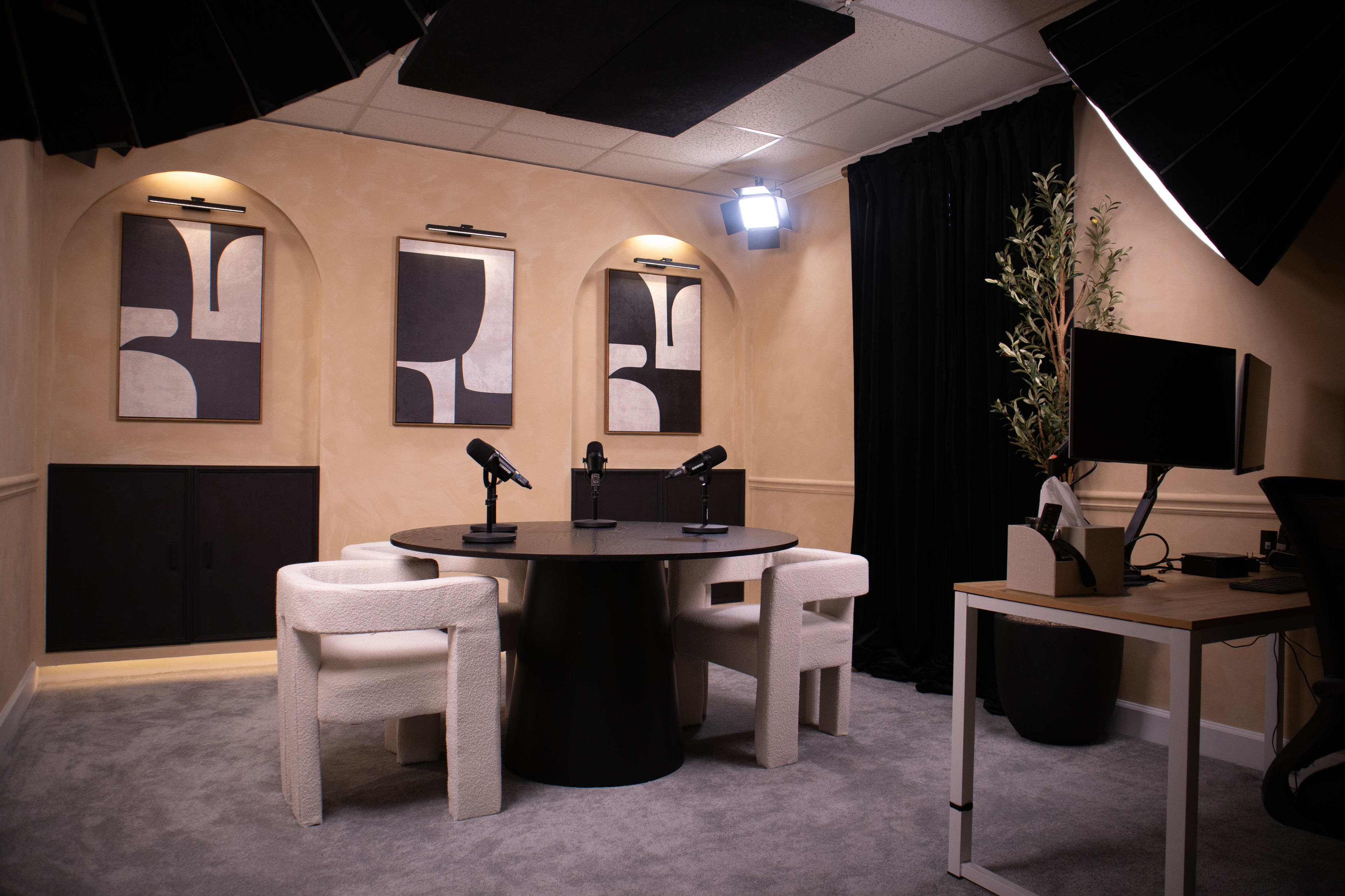 A modern recording studio featuring a round black table with two microphones, three white upholstered chairs, and wall art, set against a tan-colored backdrop.