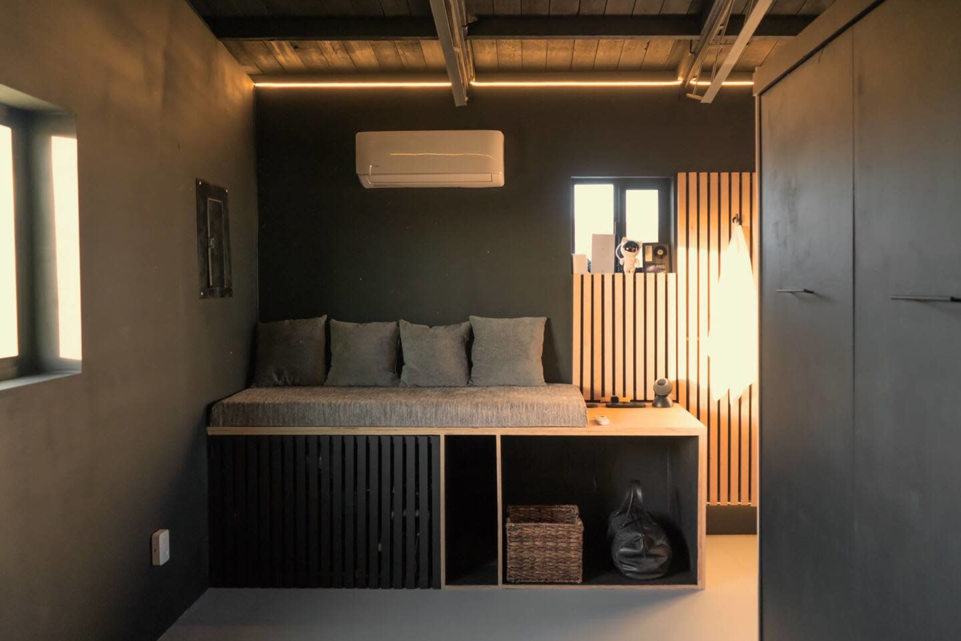 The image shows a minimalist room featuring a built-in bench with cushions, wooden accents, and an air conditioning unit on a dark wall.