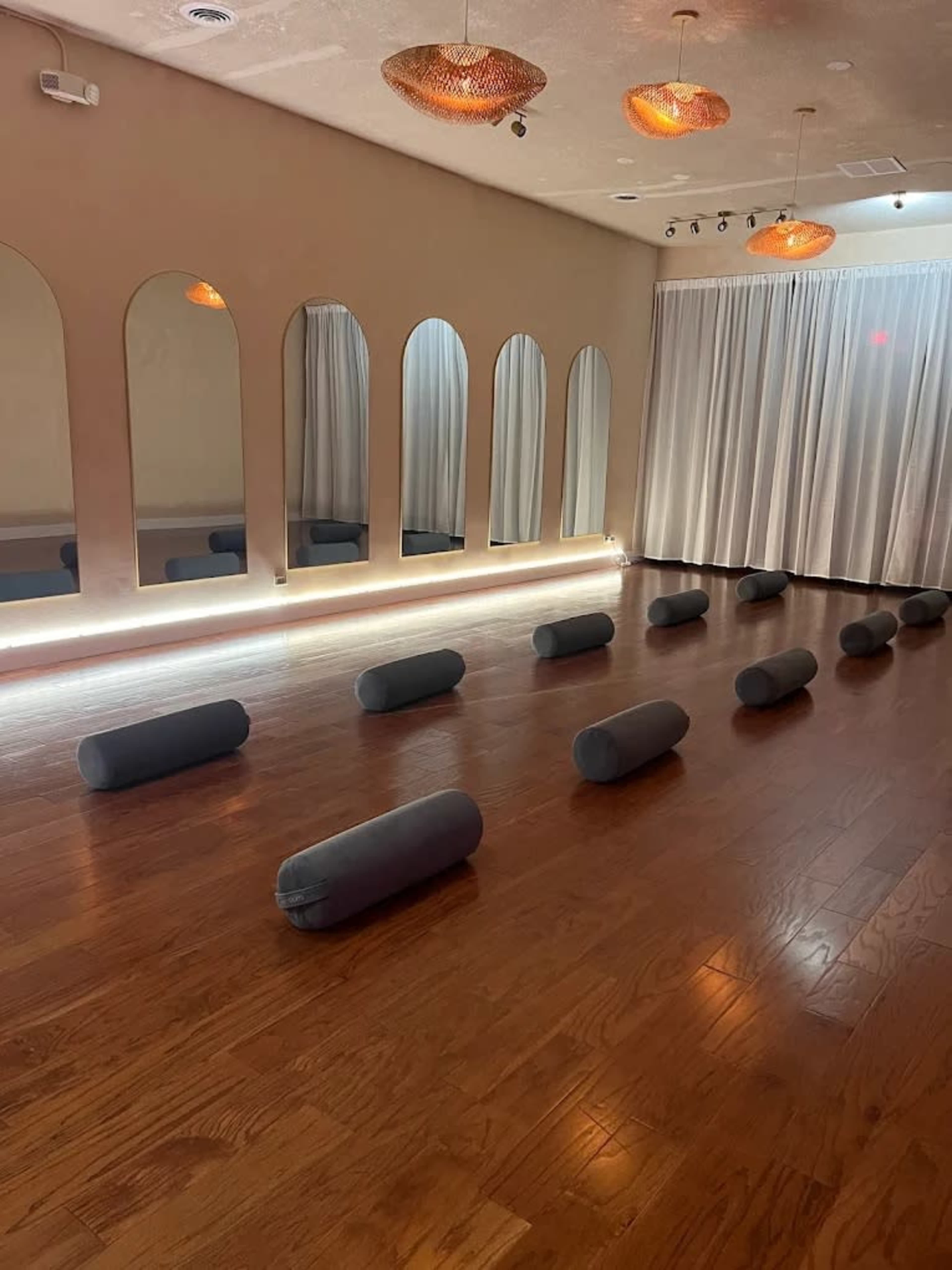 The image shows a yoga studio with a wooden floor, several gray bolsters arranged in rows, and large mirrors on one wall.