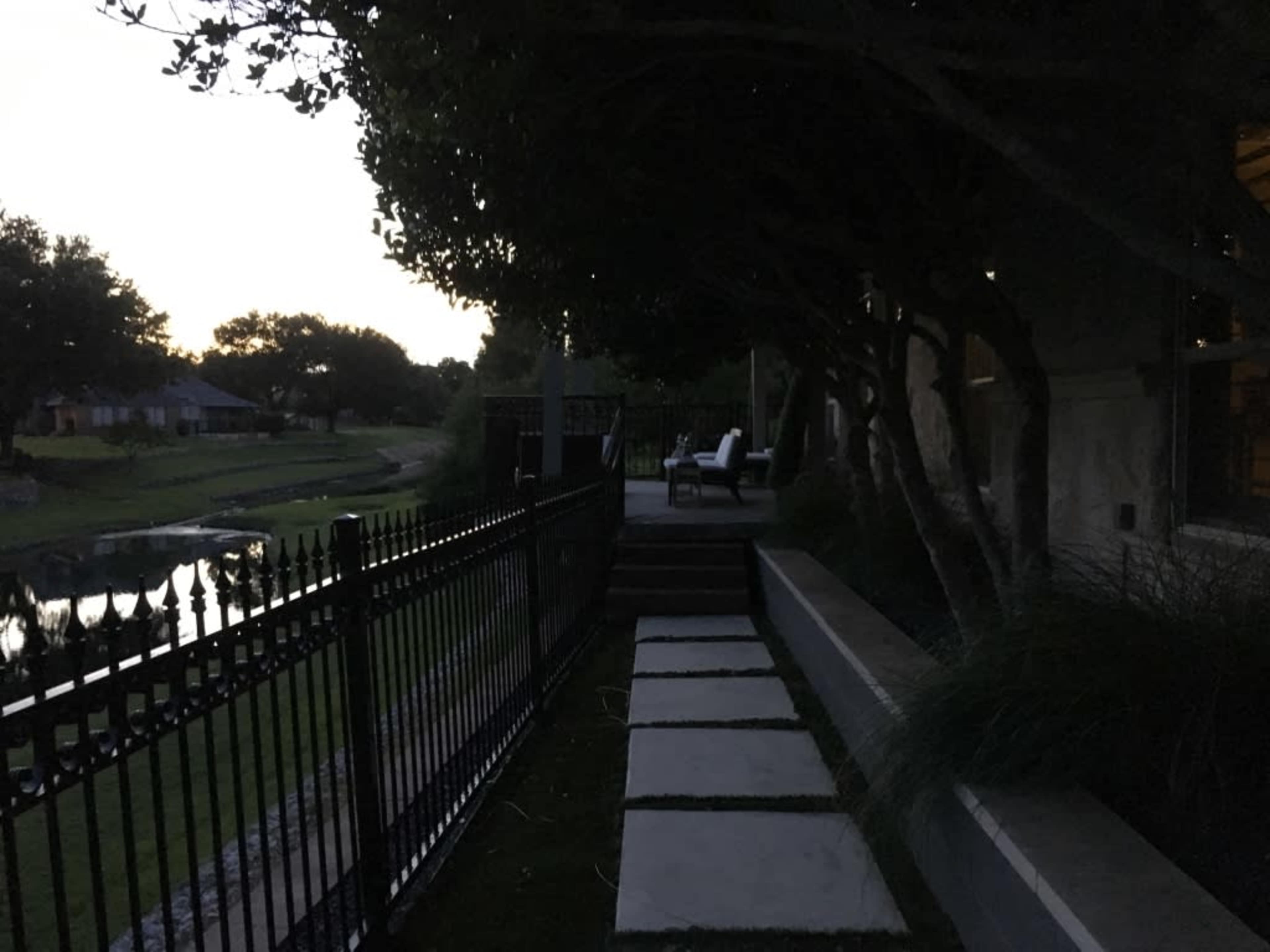 A pathway of stone slabs leads alongside a fence, with a view of a calm body of water and trees at dusk.
