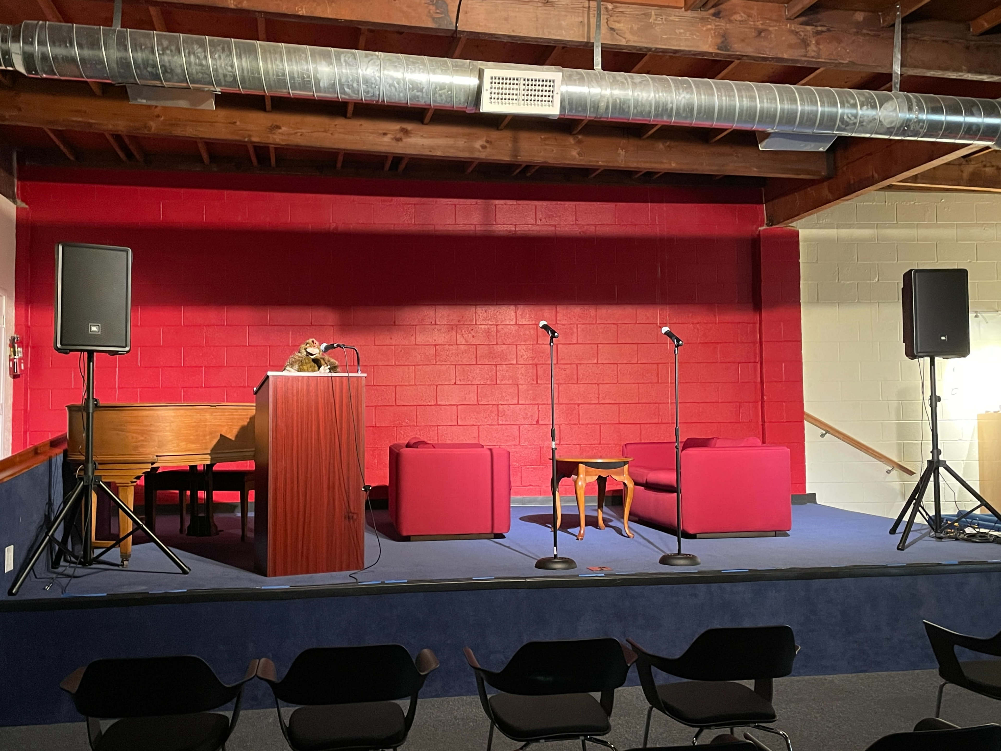 A stage setup with a podium, two microphones, a small table, and two red sofas against a red wall.