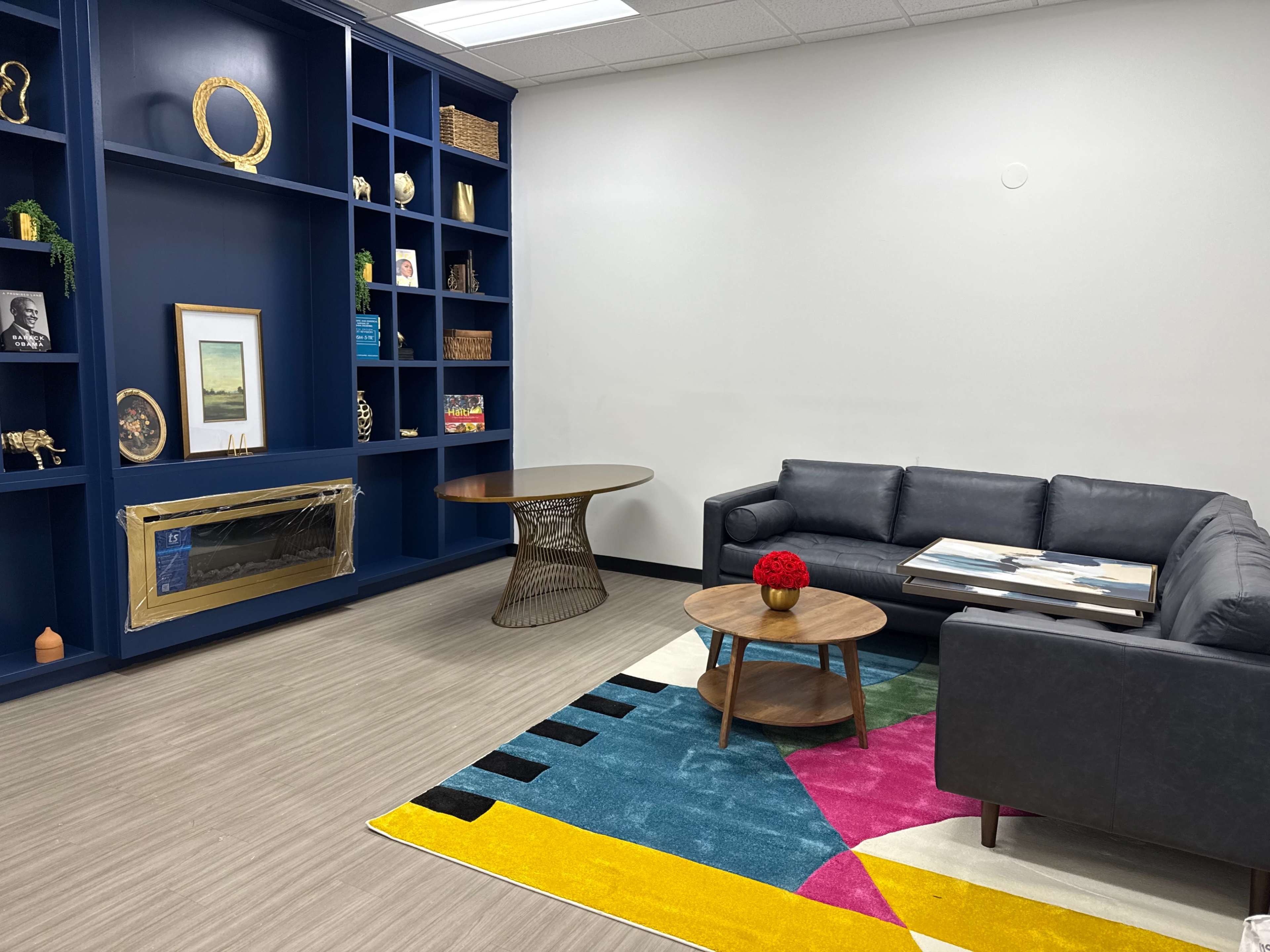The image shows a modern lounge area featuring a blue shelving unit, a gray sectional sofa, a round coffee table, and a colorful area rug.