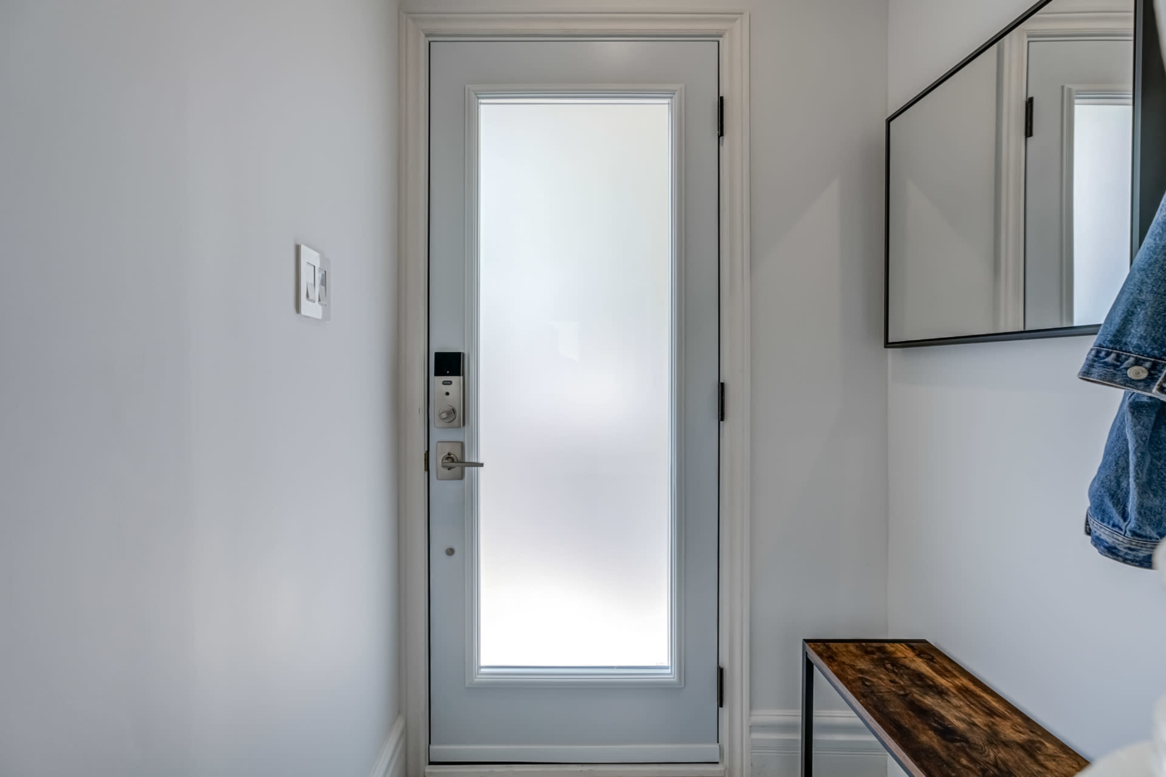 A modern entrance area with a frosted glass door, a wall-mounted mirror, and a wooden bench against a white wall.