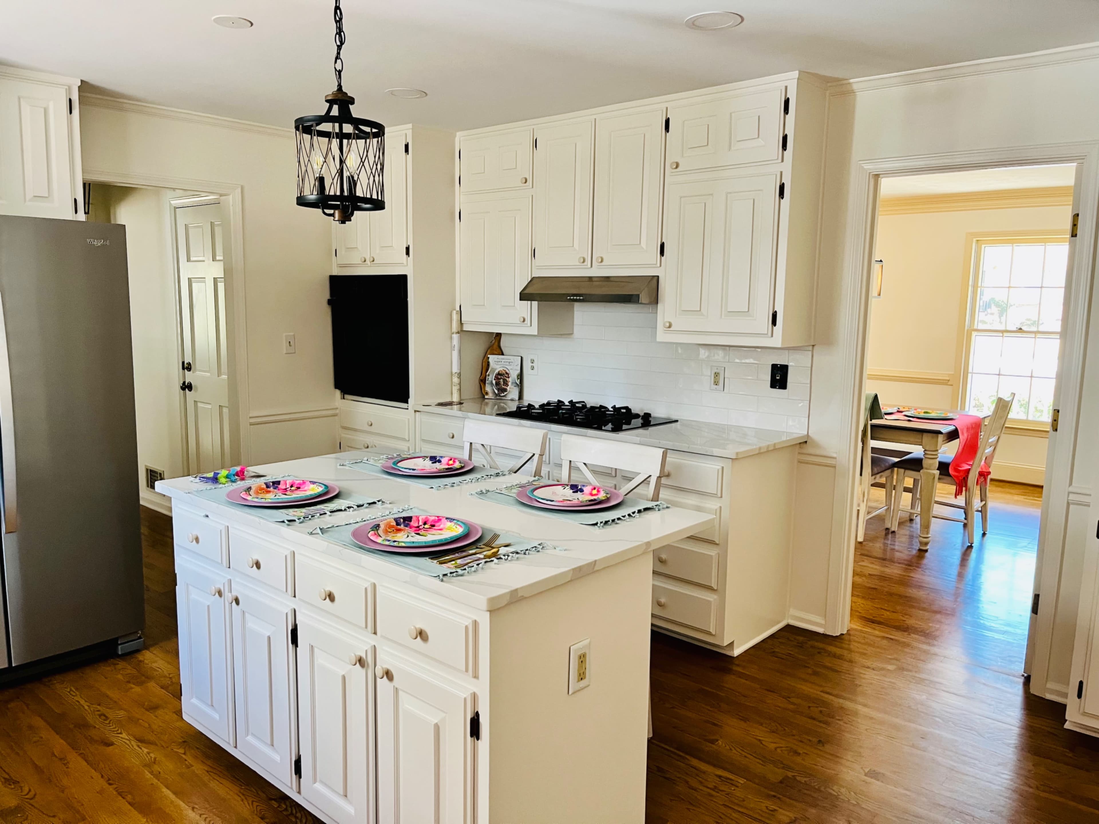 A well-lit kitchen features white cabinetry, a central island with colorful plates set for four, and an adjacent dining area visible through an open doorway.