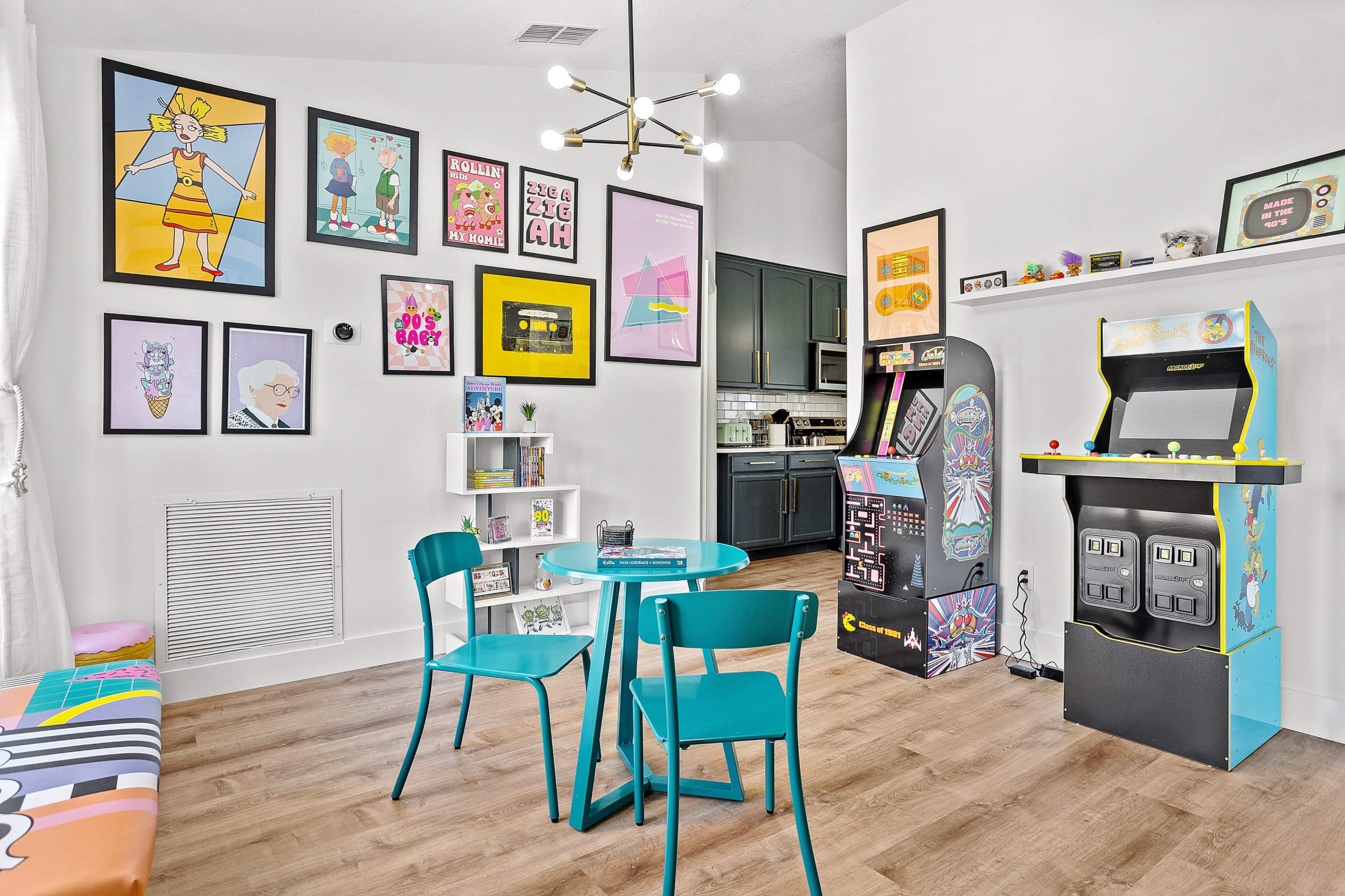 A colorful room featuring a small table with turquoise chairs, art-covered walls, and two retro arcade game machines in the corner.