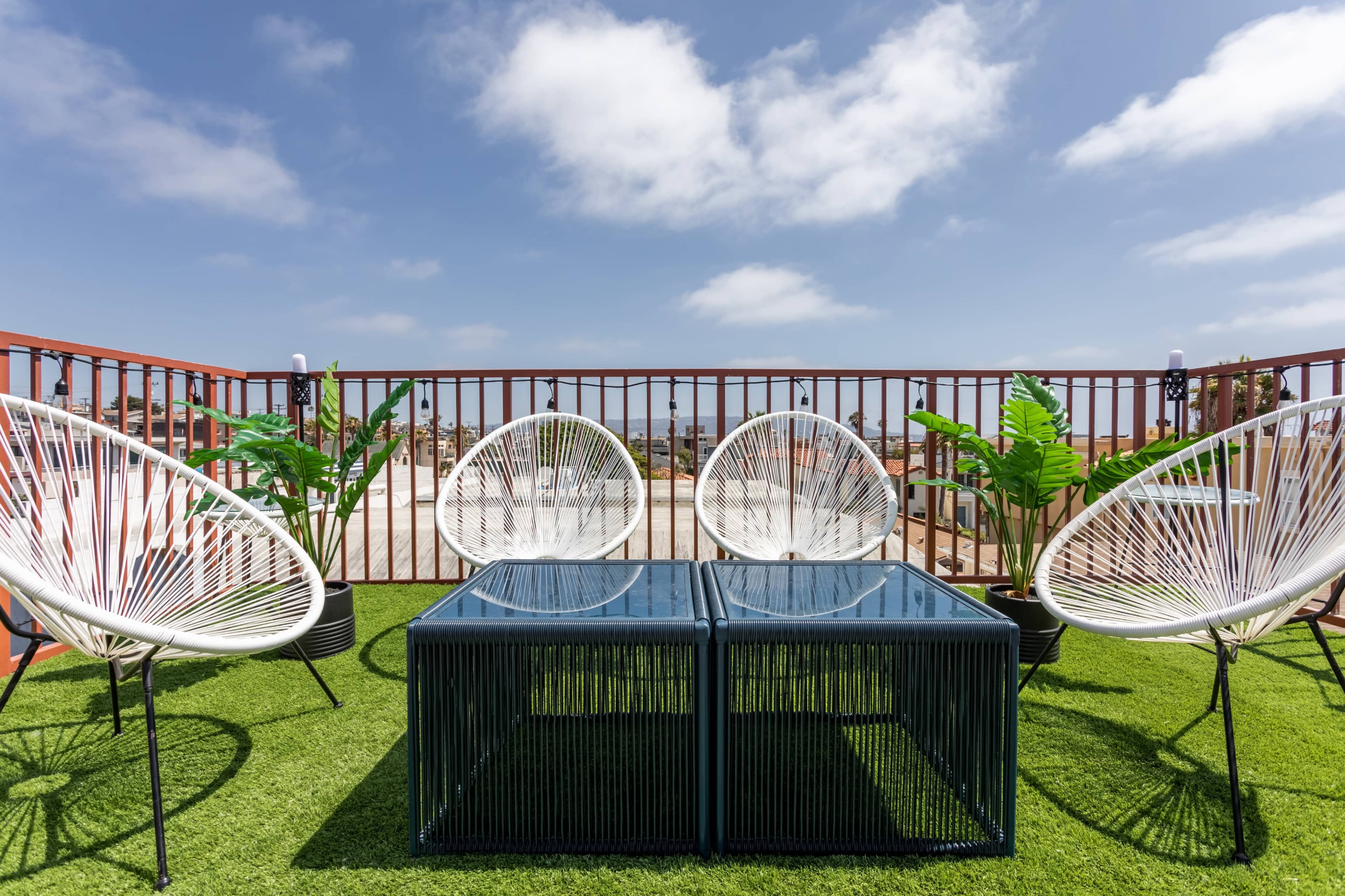 A rooftop terrace features four white chairs surrounding a glass-top table on artificial grass, with potted plants in the background.