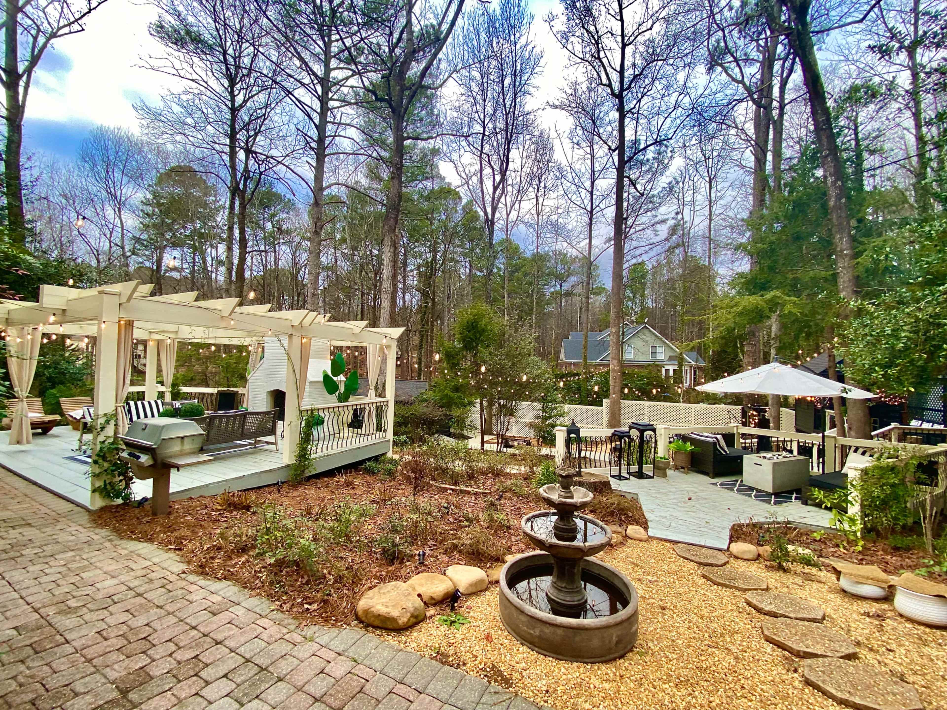 A landscaped backyard features a gazebo with seating, a stone fountain, and multiple gathering areas surrounded by trees.