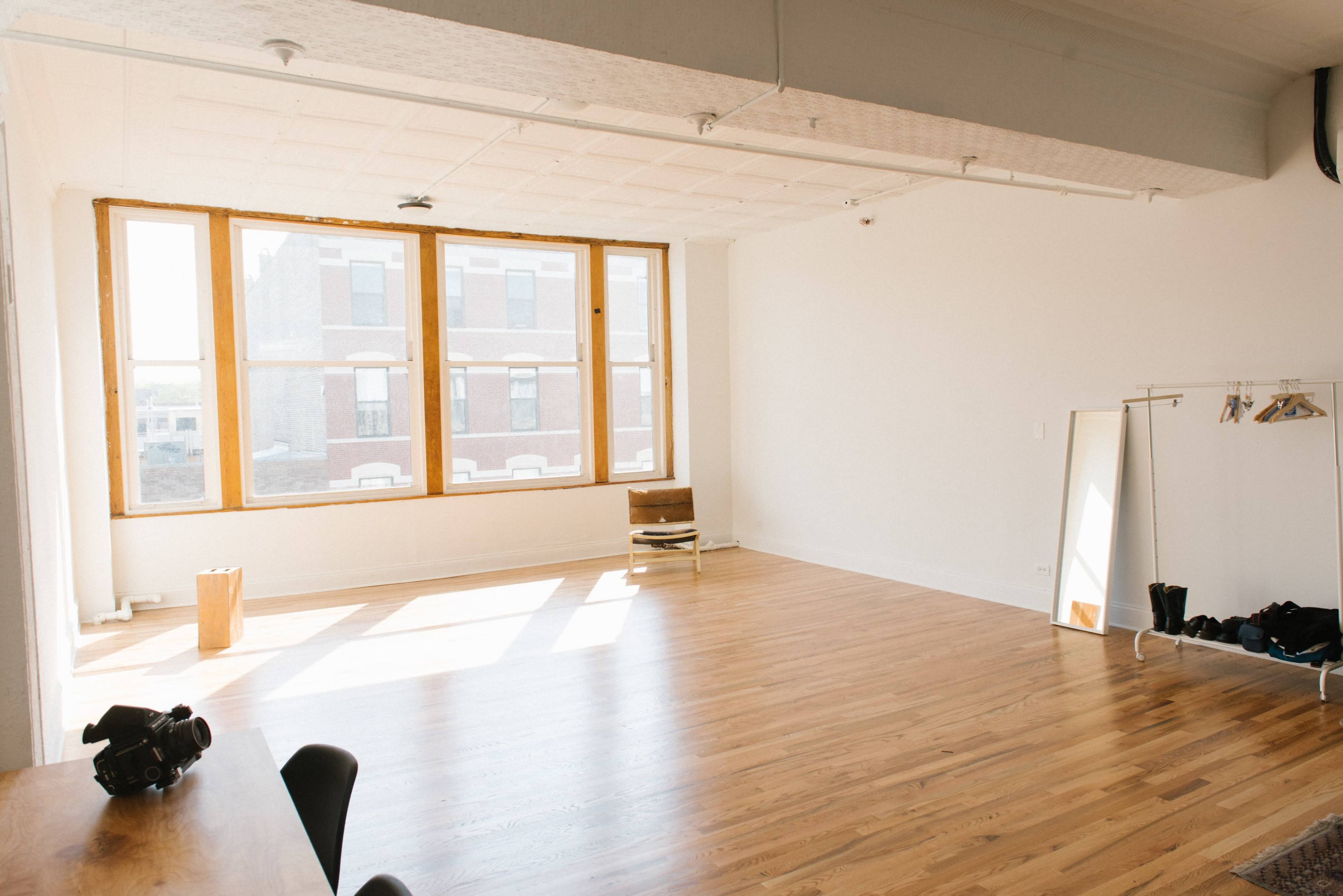 A bright, empty room features large windows, wooden flooring, and minimal furniture, including a mirror and a clothing rack.