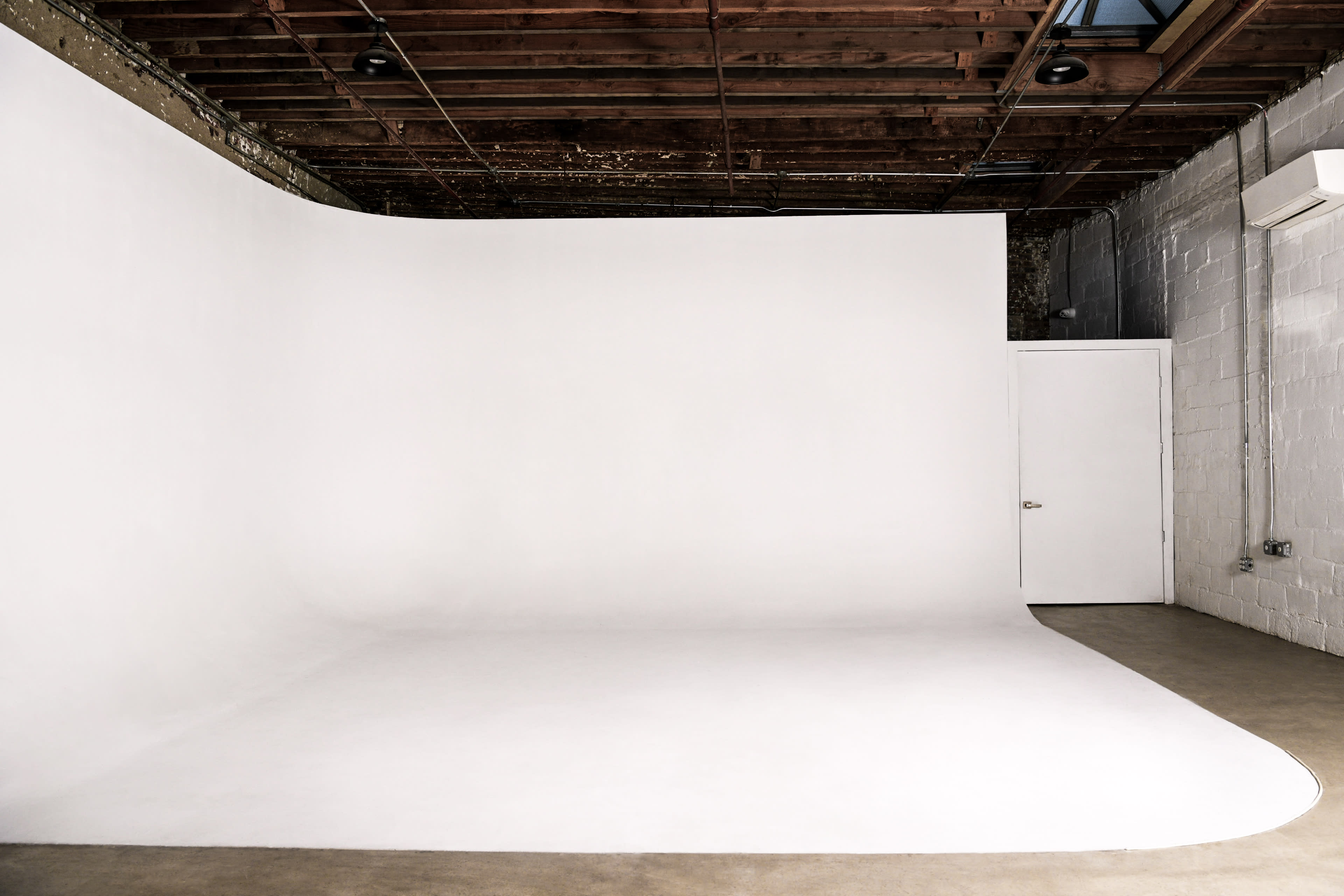 The image shows a spacious, minimalist photo studio with a seamless white backdrop and a single door against a textured wall.