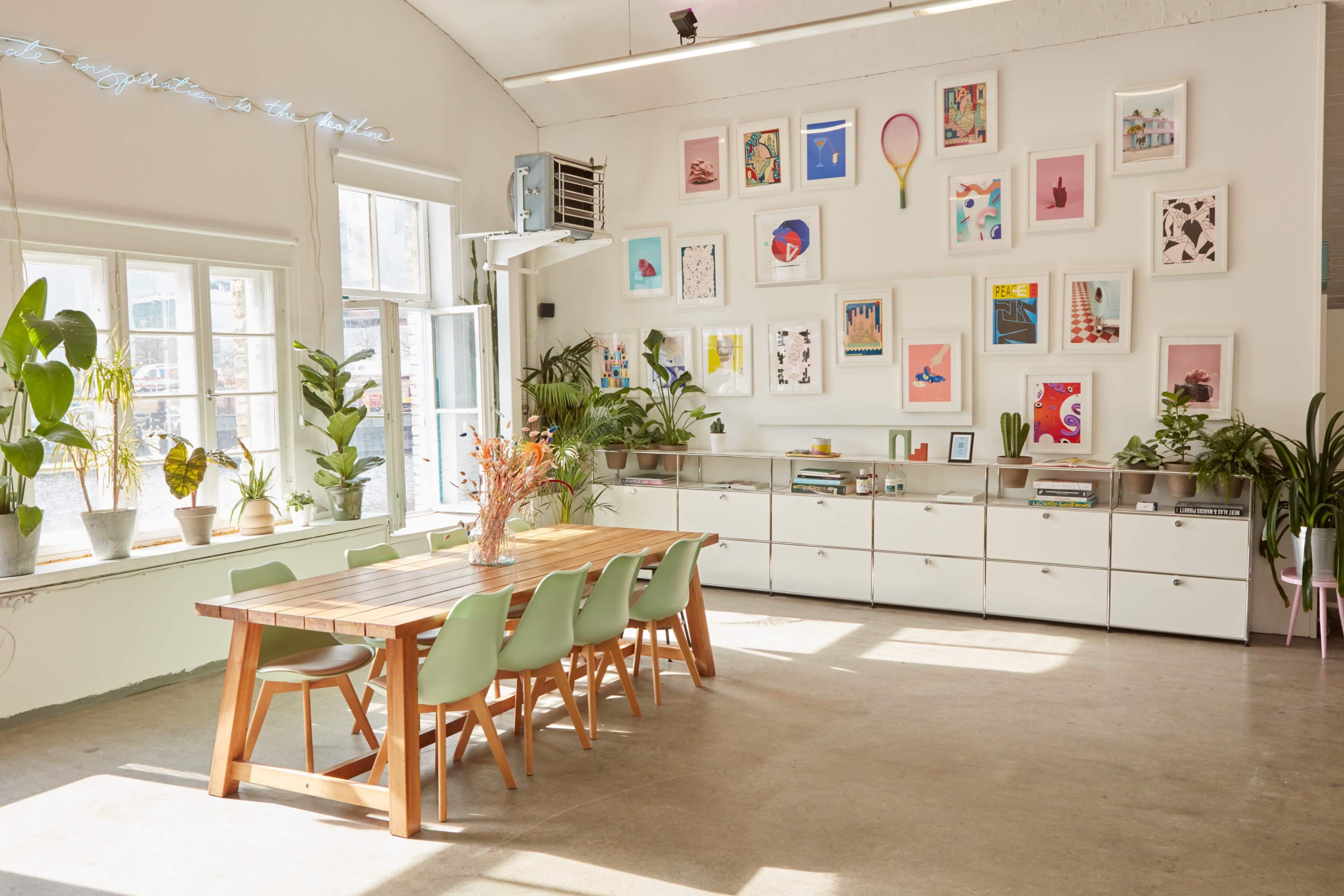 A spacious room features a large wooden table with green chairs, surrounded by plants and a wall adorned with various framed artworks.