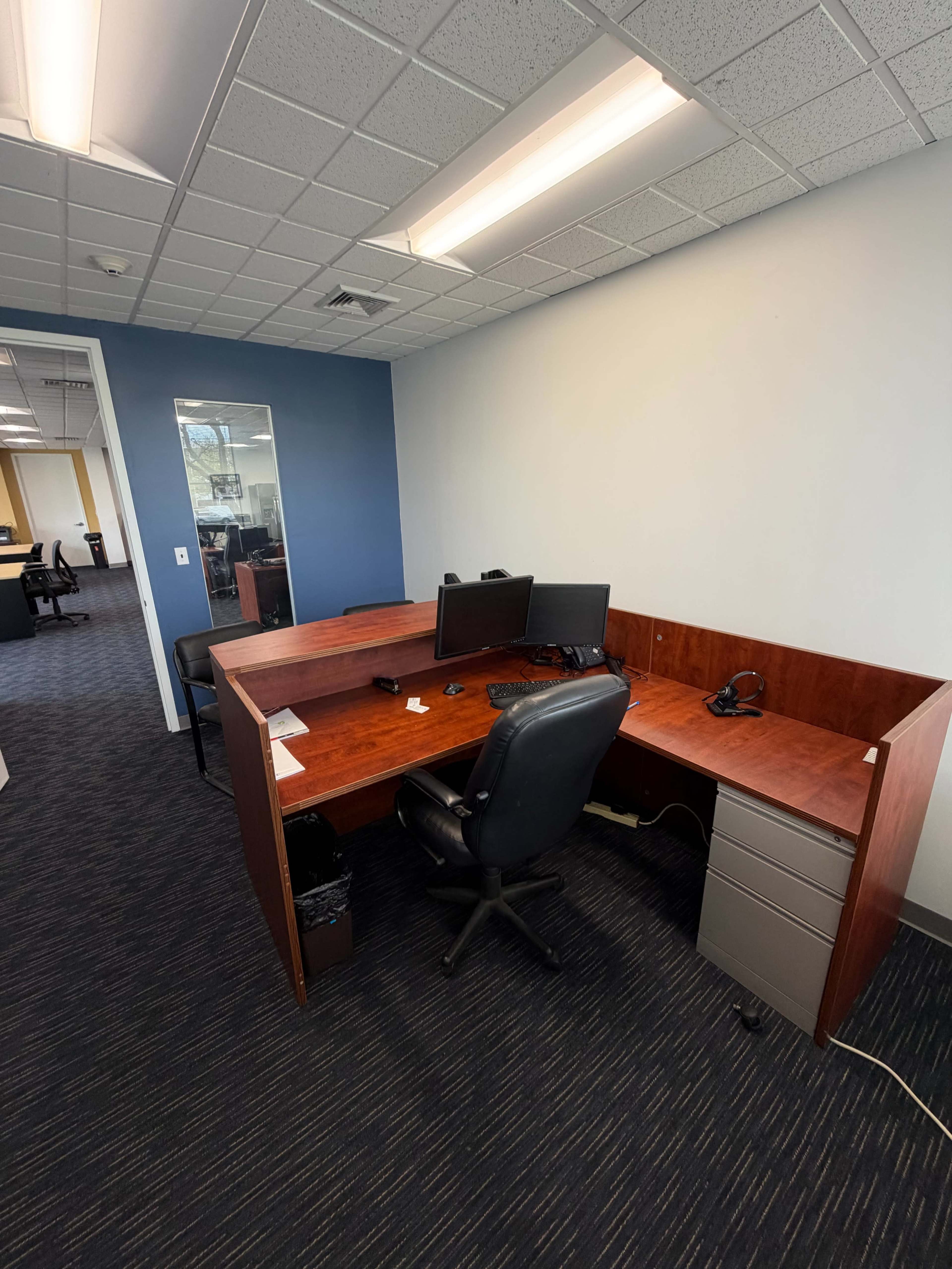 The image shows an office space featuring a wooden desk with two computer monitors, a chair, and a filing cabinet.