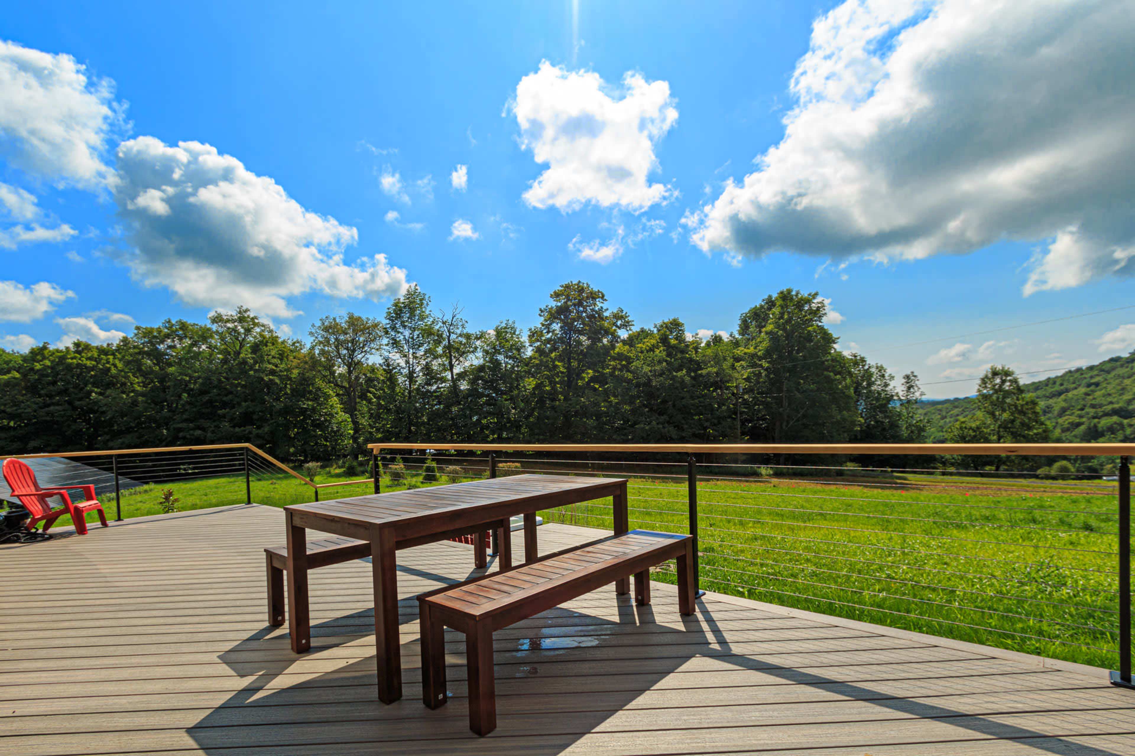 A wooden table and benches sit on a deck overlooking a green landscape with trees under a partly cloudy sky.