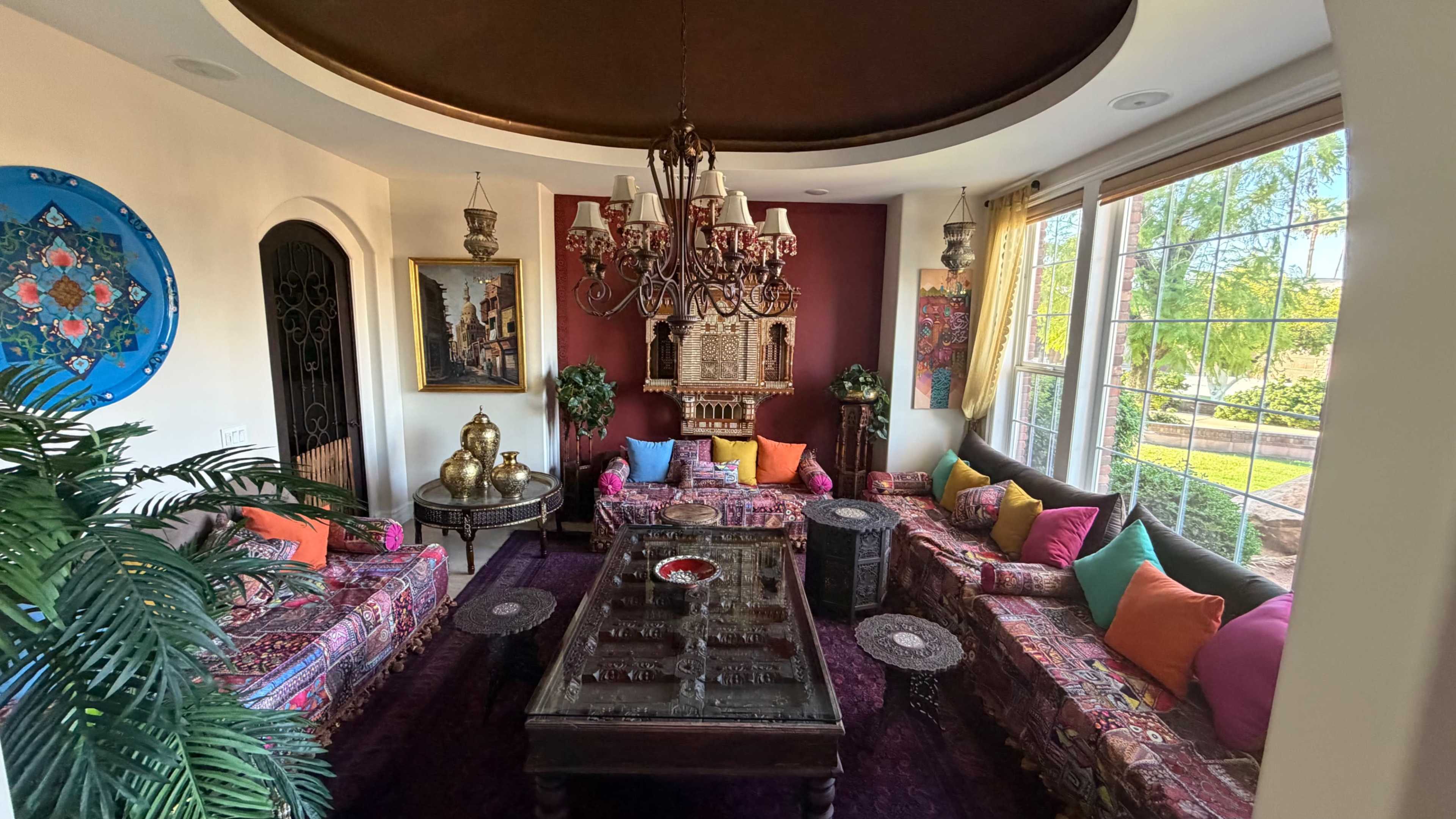 The image depicts a cozy living room with colorful upholstered sofas, a chandelier, and large windows revealing a garden view.
