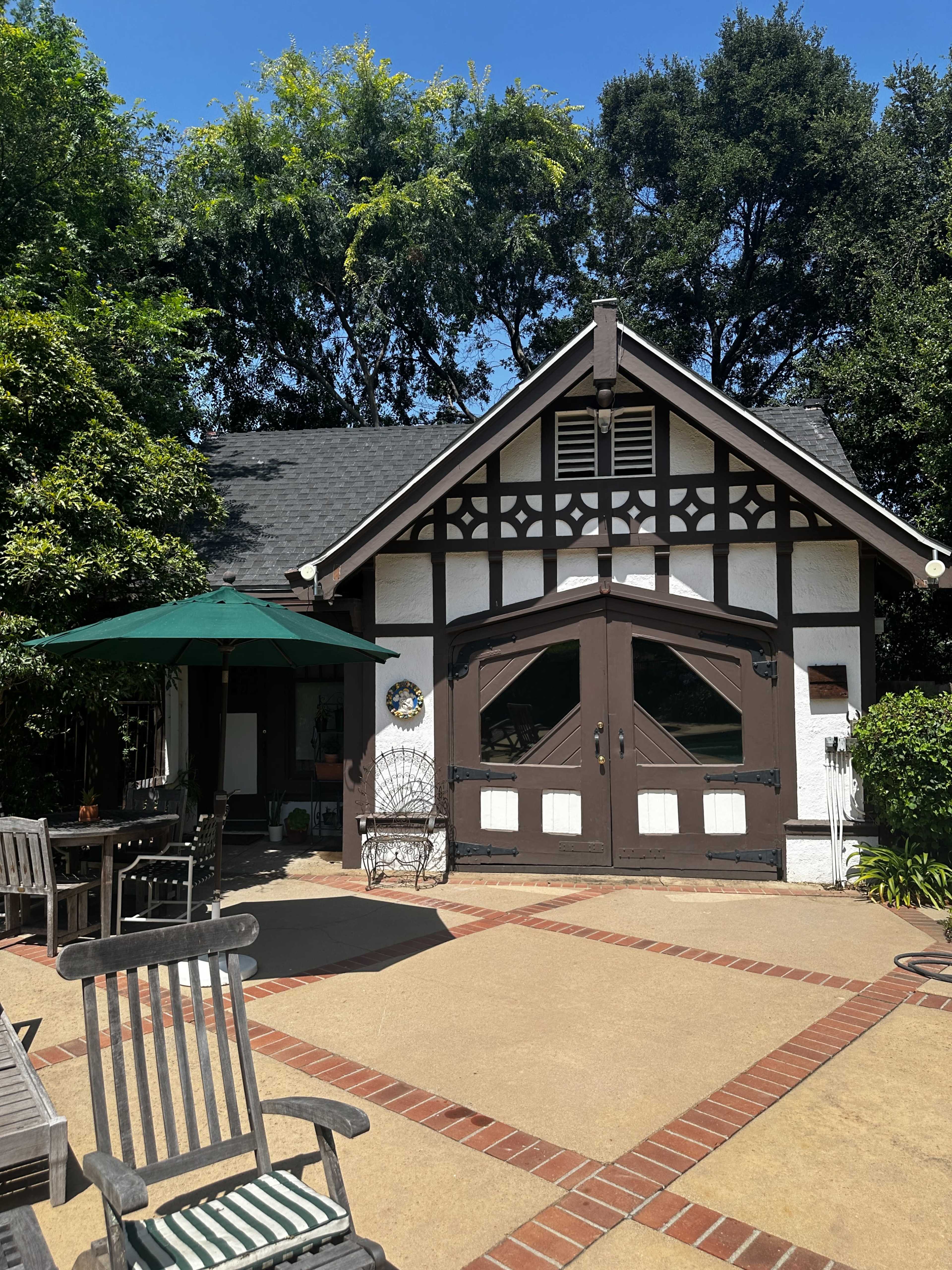 A small, charming cottage with a decorative facade and double doors is surrounded by greenery and outdoor seating on a sunlit patio.