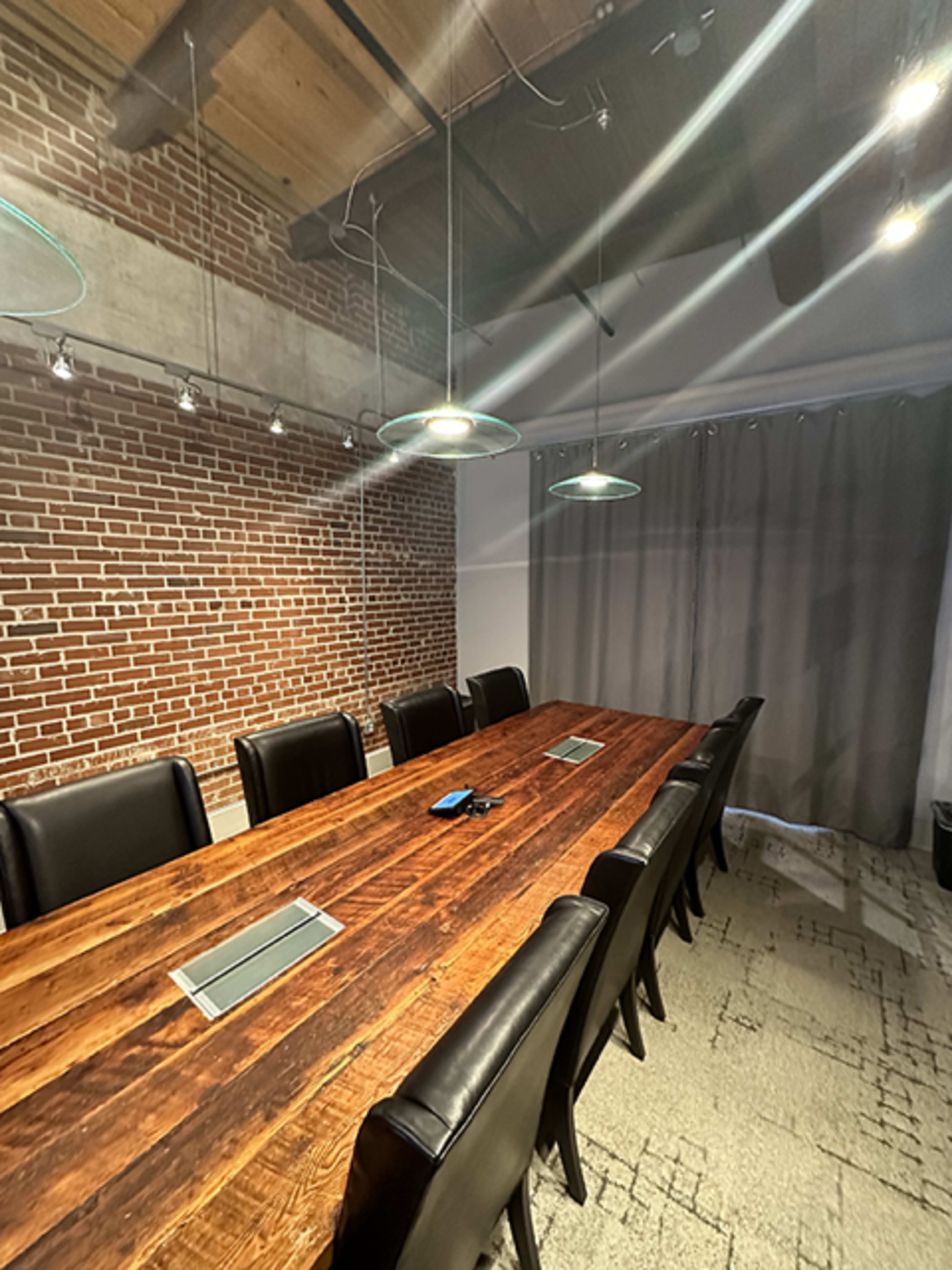 A long wooden table surrounded by black chairs is set in a conference room with exposed brick walls and overhead pendant lights.