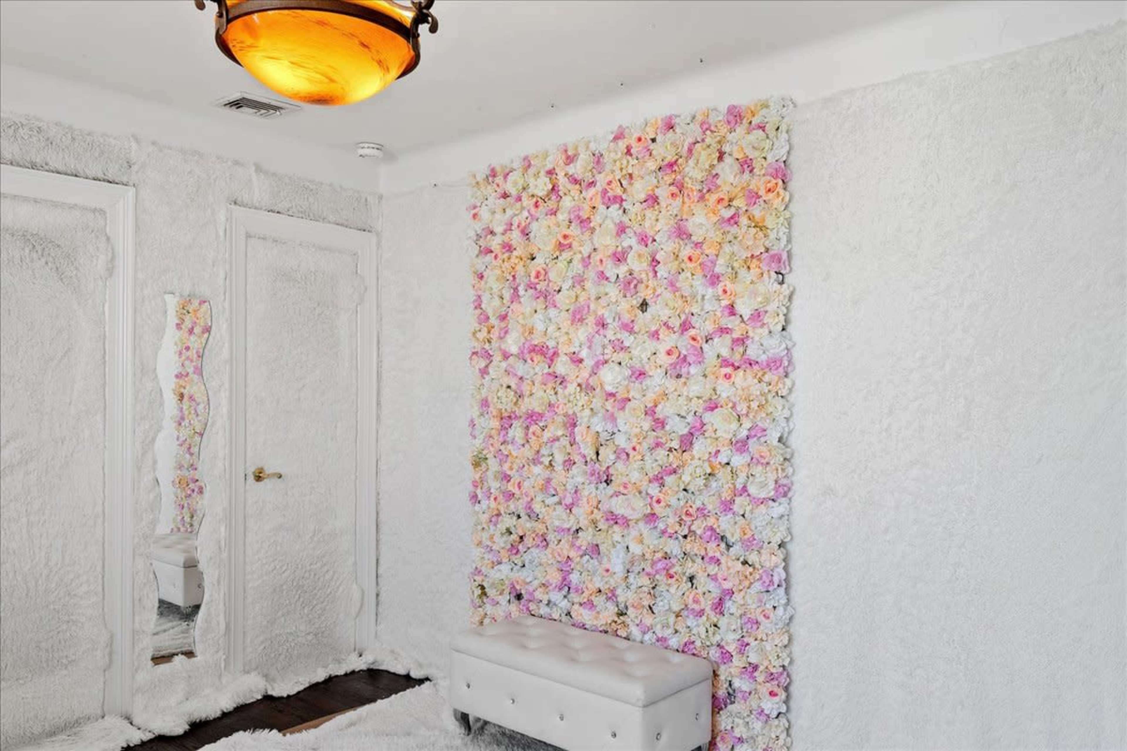 A brightly lit room with a wall covered in a floral arrangement of pink and white flowers, featuring a white tufted bench and a mirror against a soft, textured backdrop.