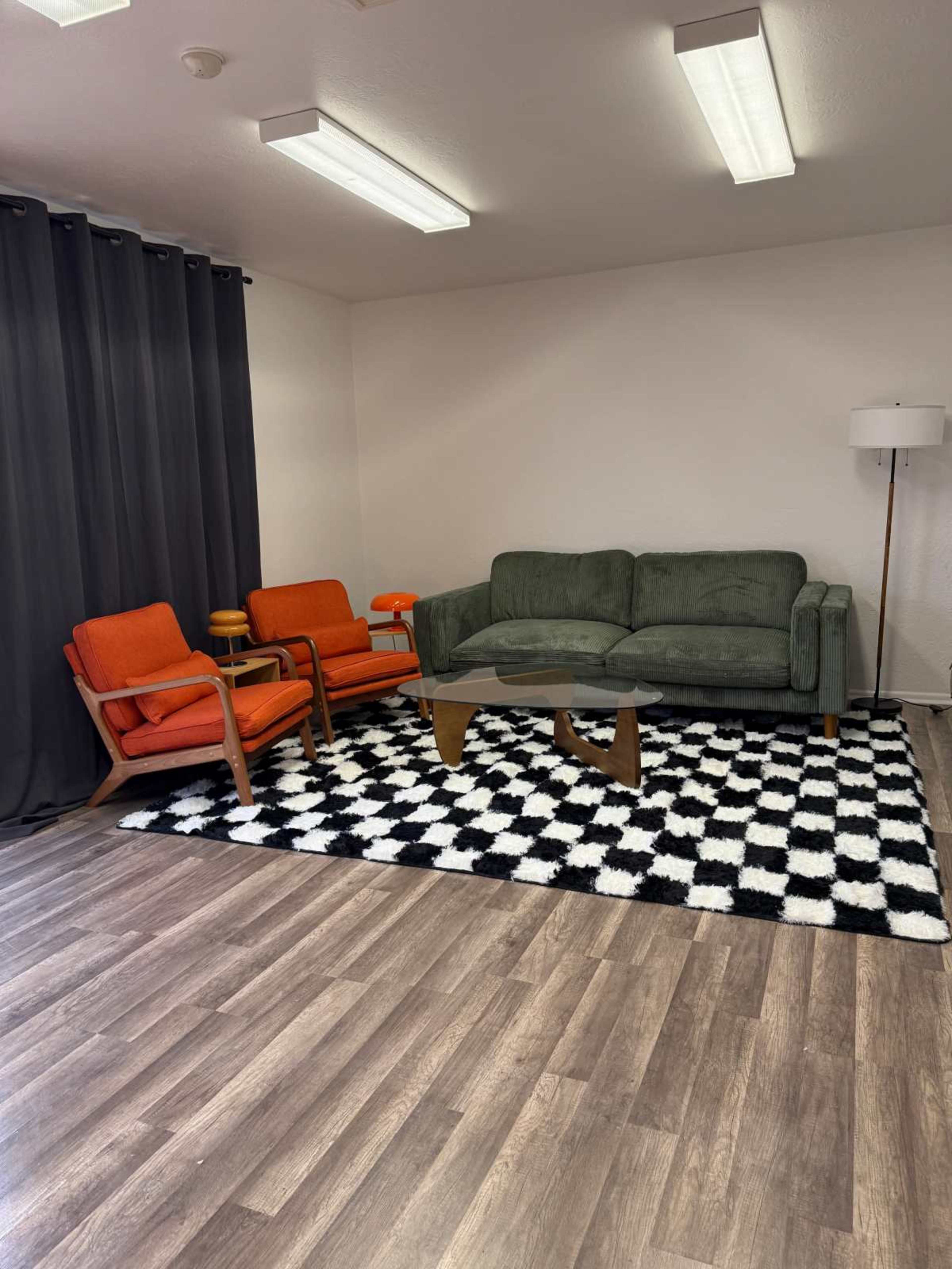 The image shows a small living space featuring a green sofa, two orange armchairs, a glass coffee table, and a black-and-white checkered rug on a wooden floor, with dark curtains in the background.