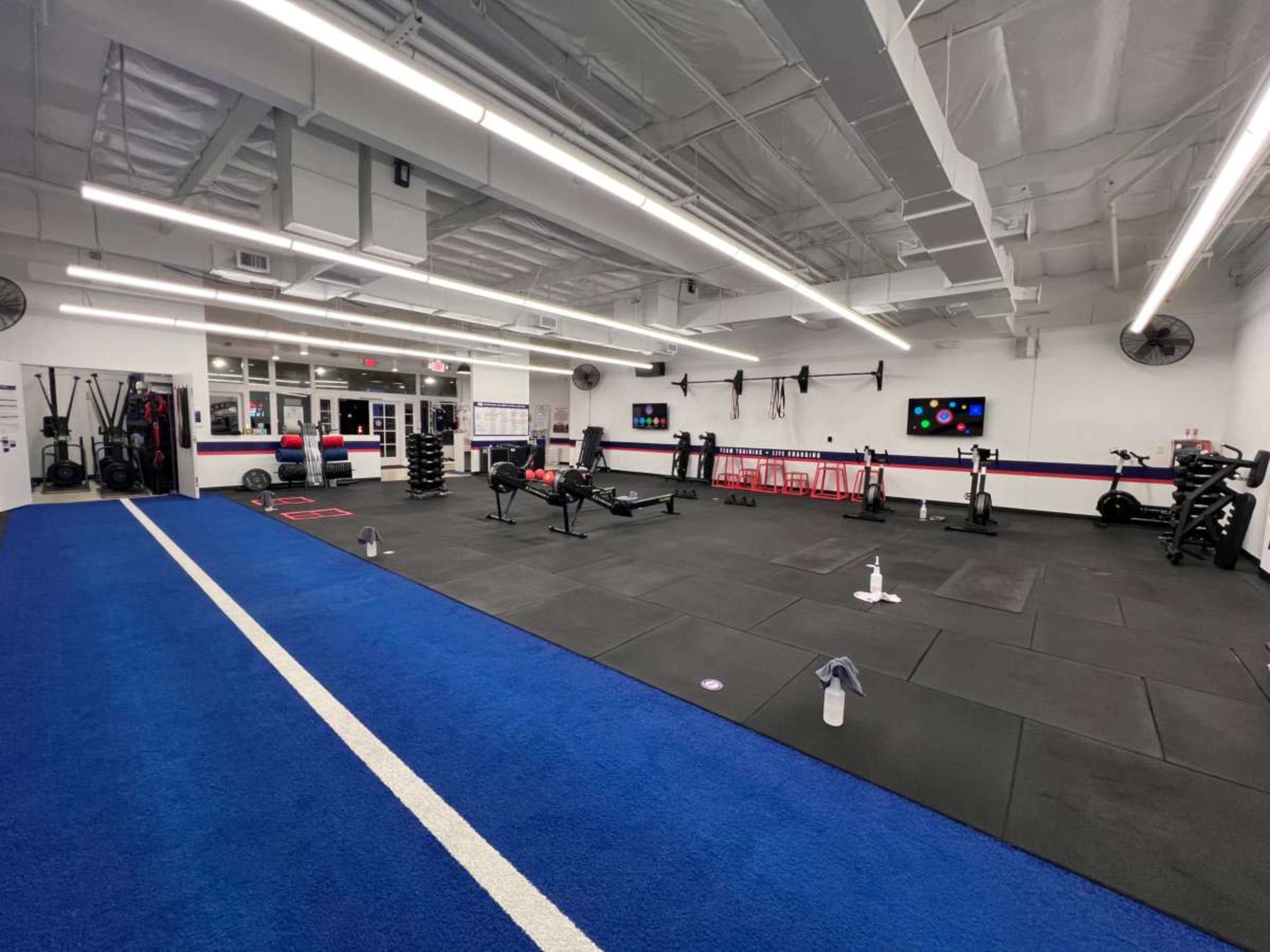 The image shows a modern gym with exercise equipment arranged on rubber flooring, featuring weightlifting stations, cardio machines, and a blue strip running along one side.
