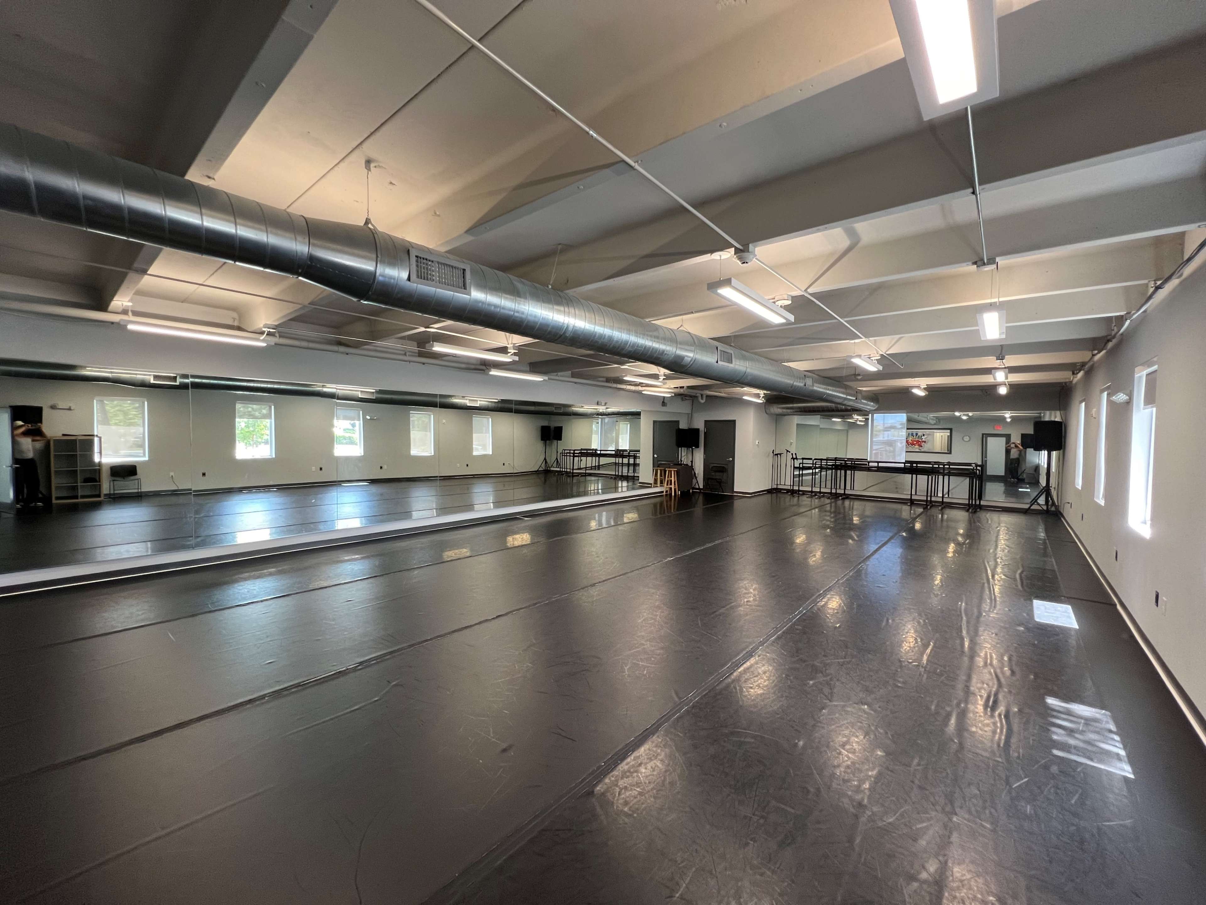 A spacious dance studio with a black, glossy floor, large mirrors along one wall, and windows providing natural light.
