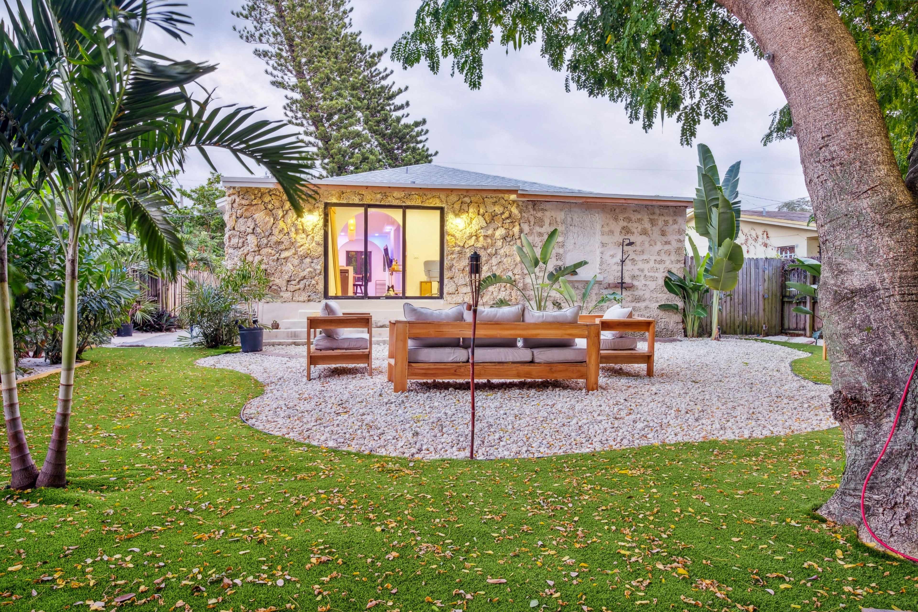 The image shows a landscaped backyard with a stone house in the background, featuring a wooden seating area on a pebbled surface surrounded by green grass and tropical plants.