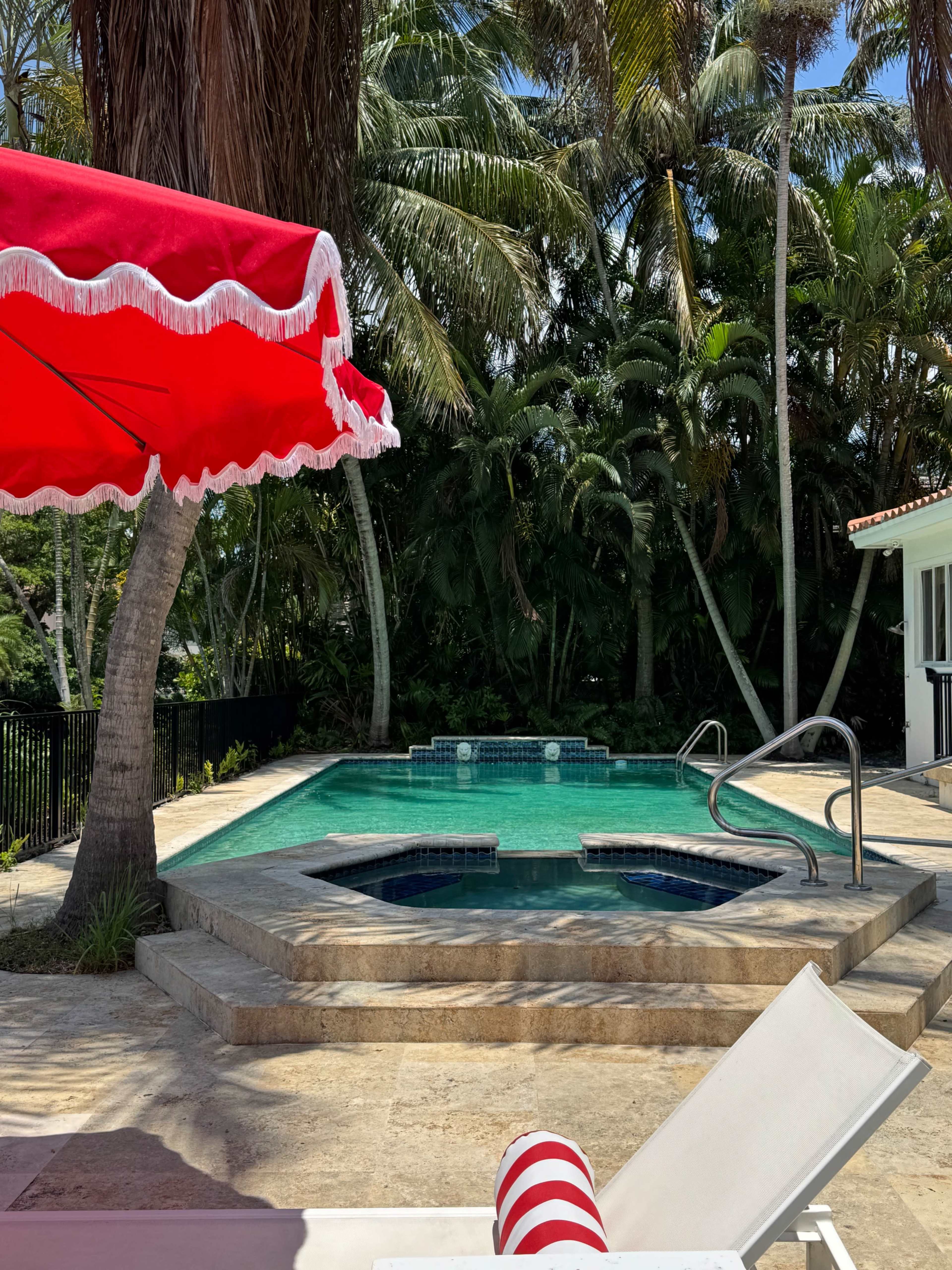 The image shows a tropical outdoor pool area with a large red umbrella, a Jacuzzi, and lush greenery surrounding the swimming pool.