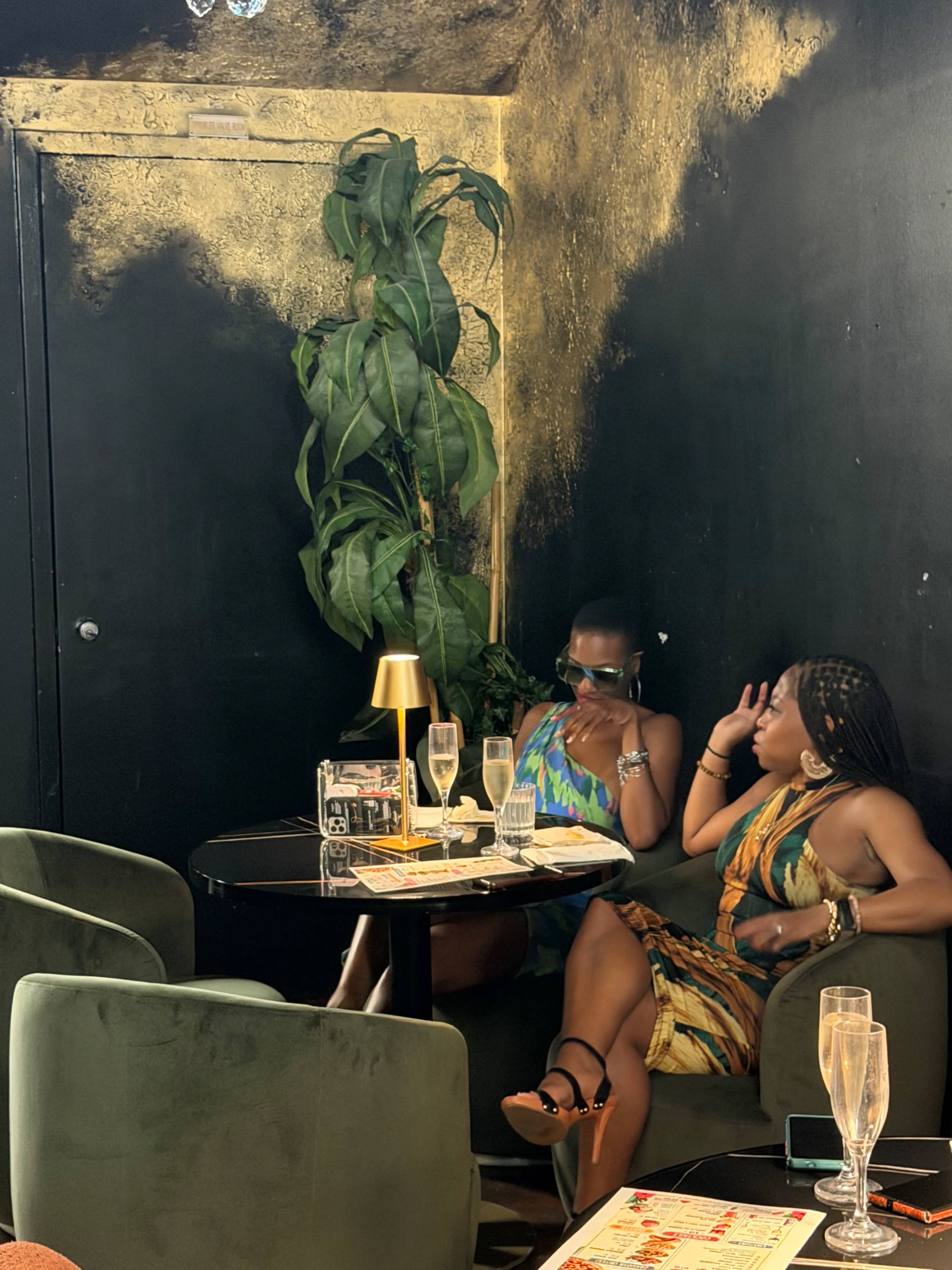 Two women are seated at a small table in a stylish, dimly lit restaurant, surrounded by dark walls and lush greenery.