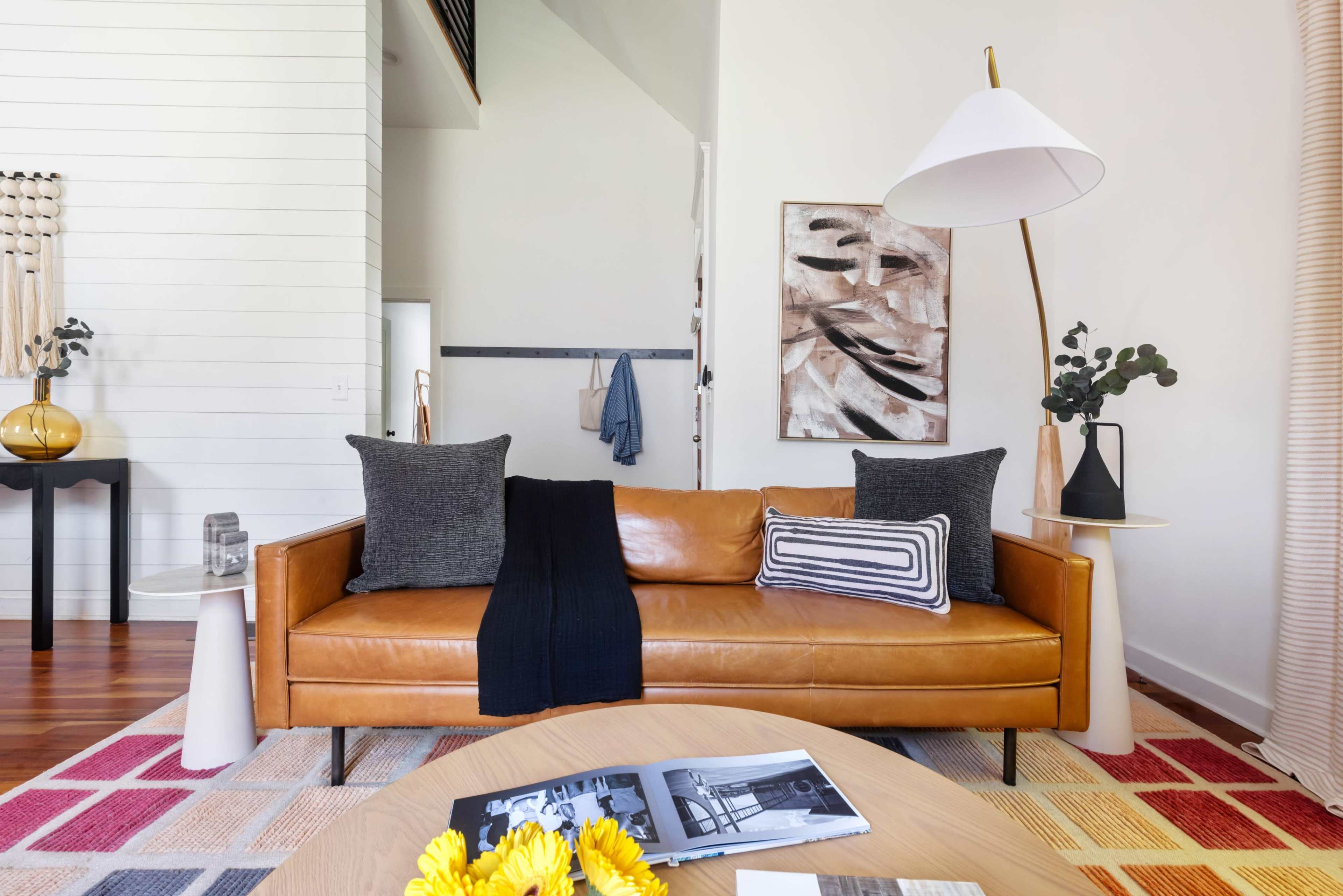 A brown leather sofa with decorative pillows is positioned in a bright, modern living room featuring a coffee table and artwork on the walls.