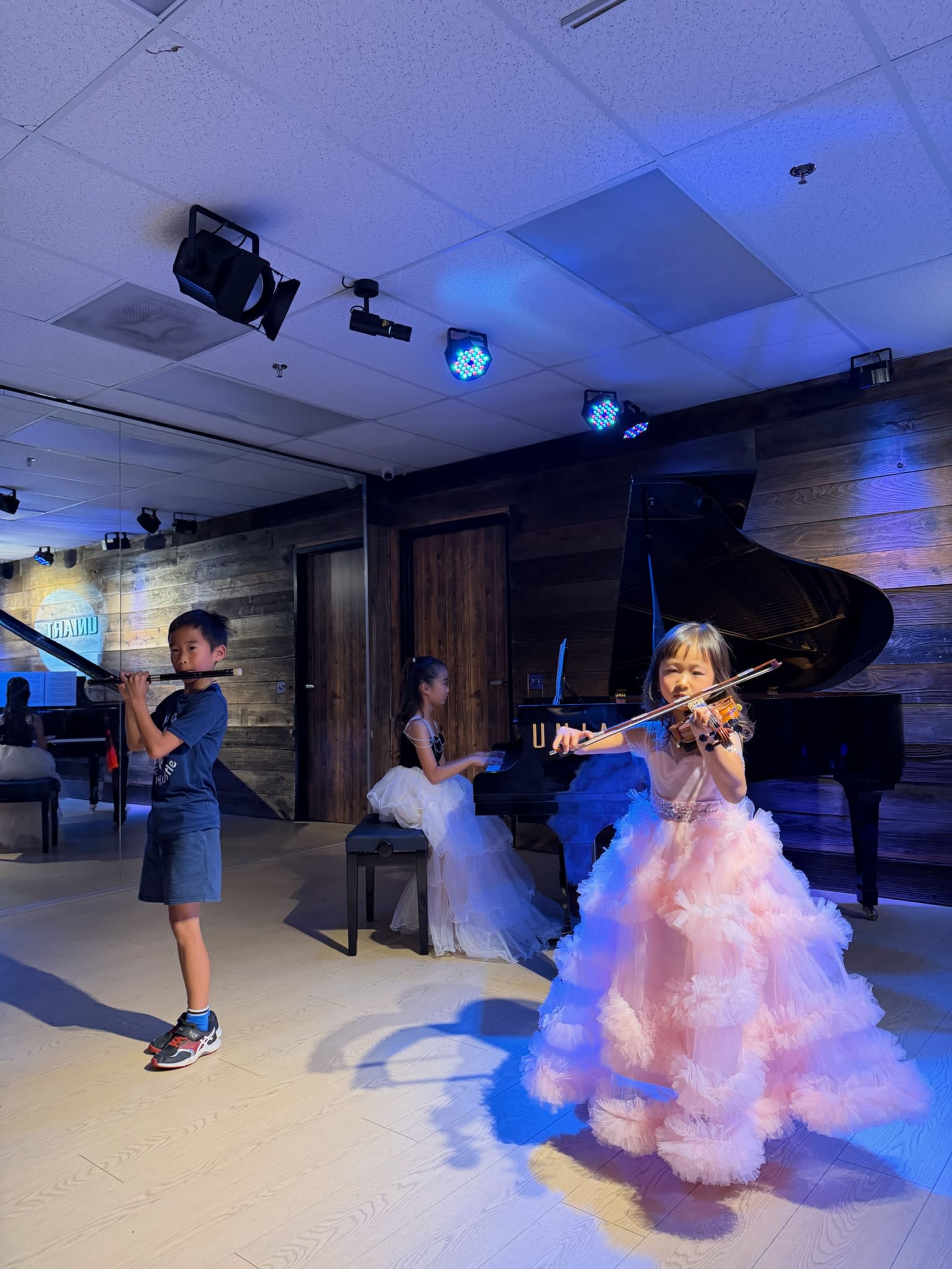 A young girl in a pink dress plays the violin, while a boy in a blue outfit plays the flute and another girl sits at a piano in a music studio with blue lights.