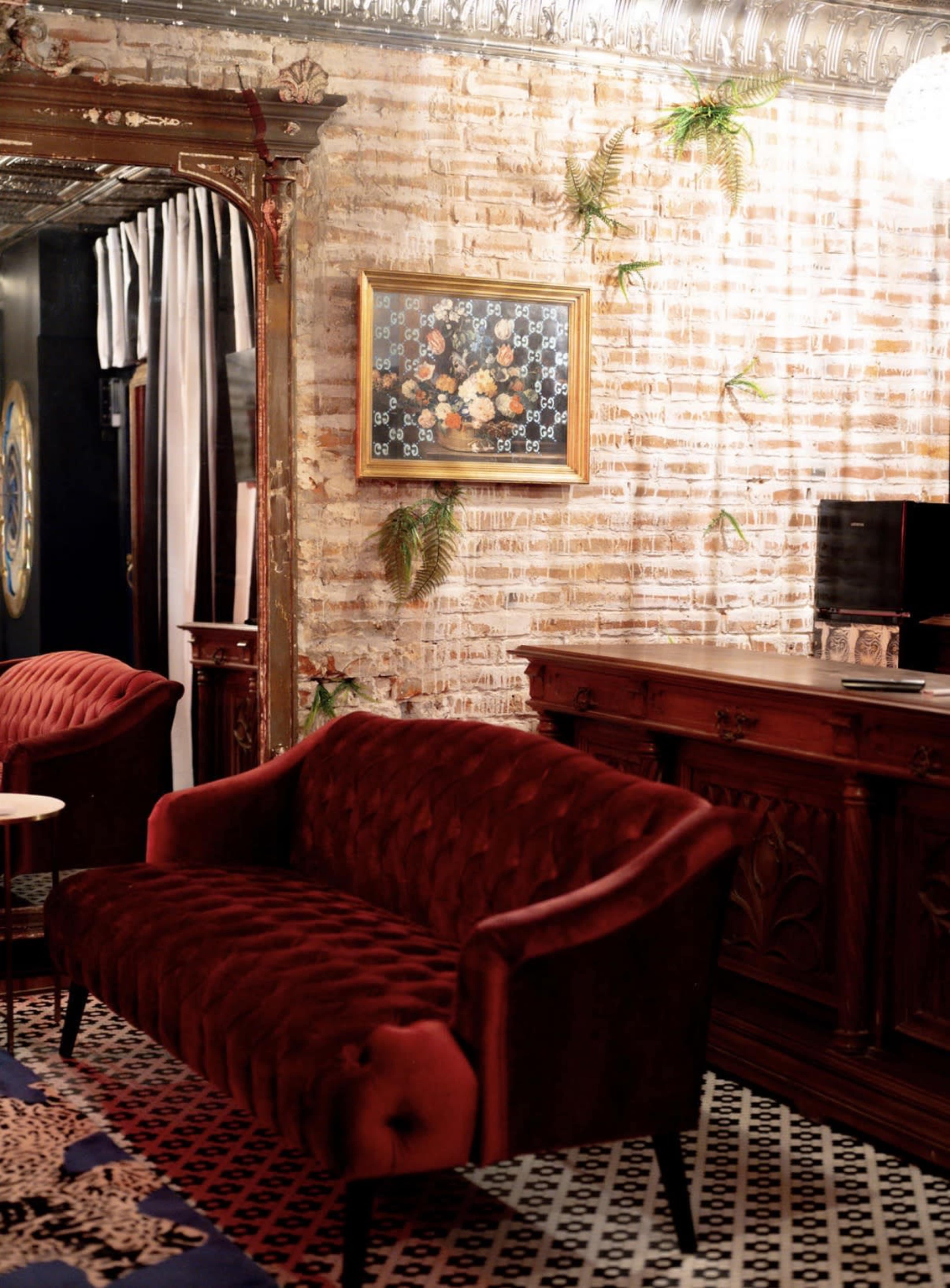 The image shows a cozy sitting area with a red tufted velvet sofa, a dark wood sideboard, and a decorative floral painting hanging on a brick wall.
