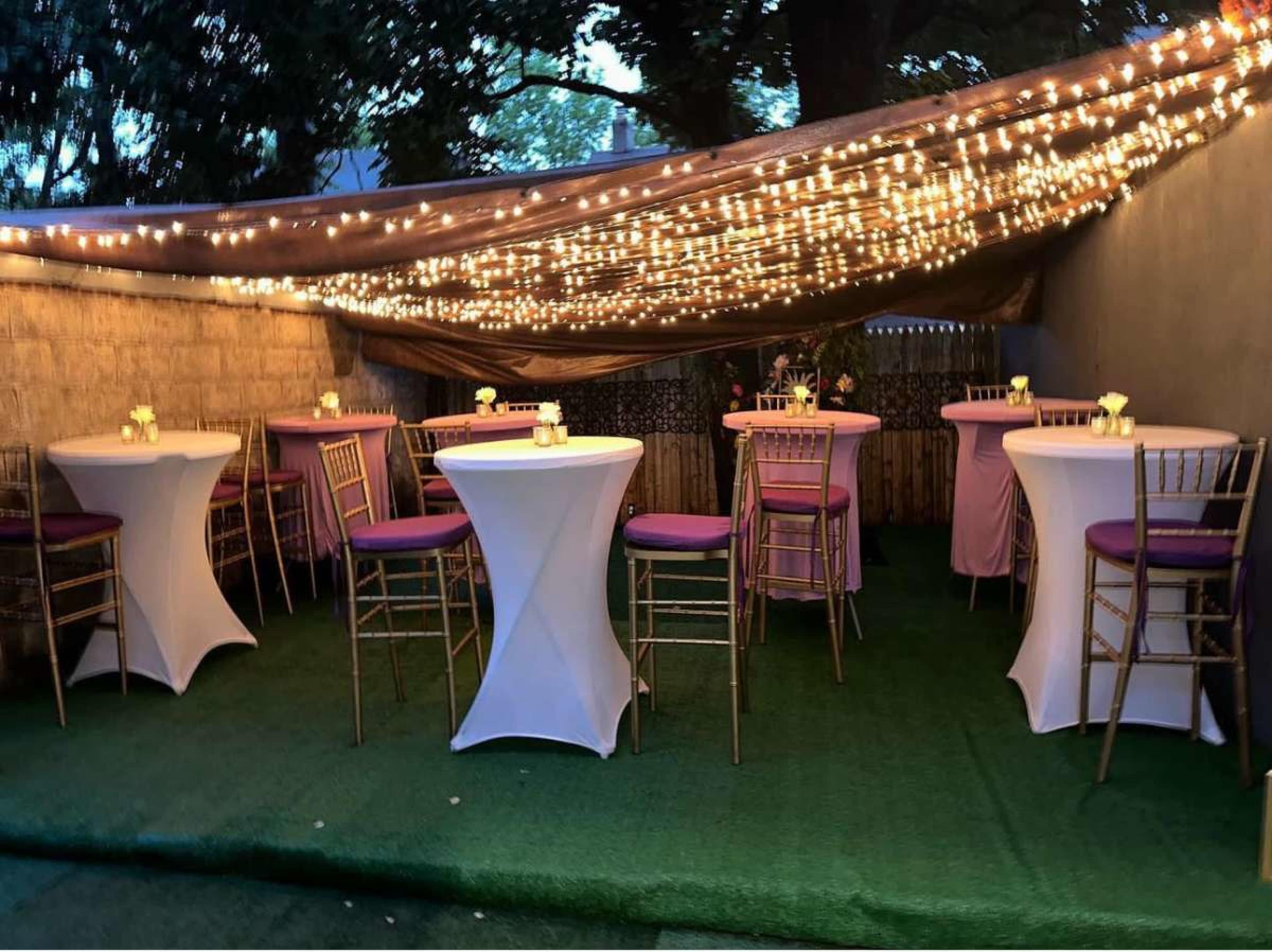 A decorated outdoor space features white cocktail tables with purple seating under a canopy of string lights.