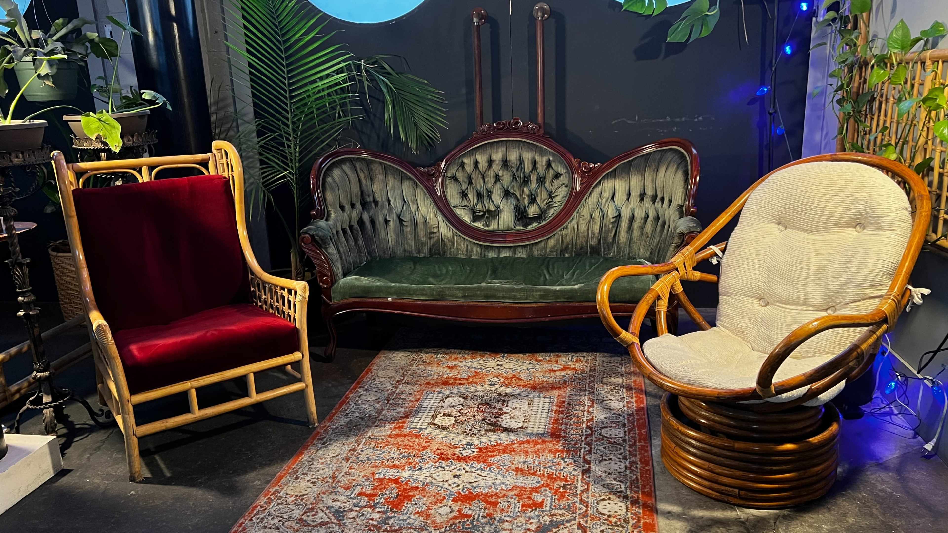 A cozy seating arrangement with a vintage green upholstered sofa, a red velvet wingback chair, and a circular rattan chair, all placed on a patterned area rug against a backdrop of plants and soft lighting.