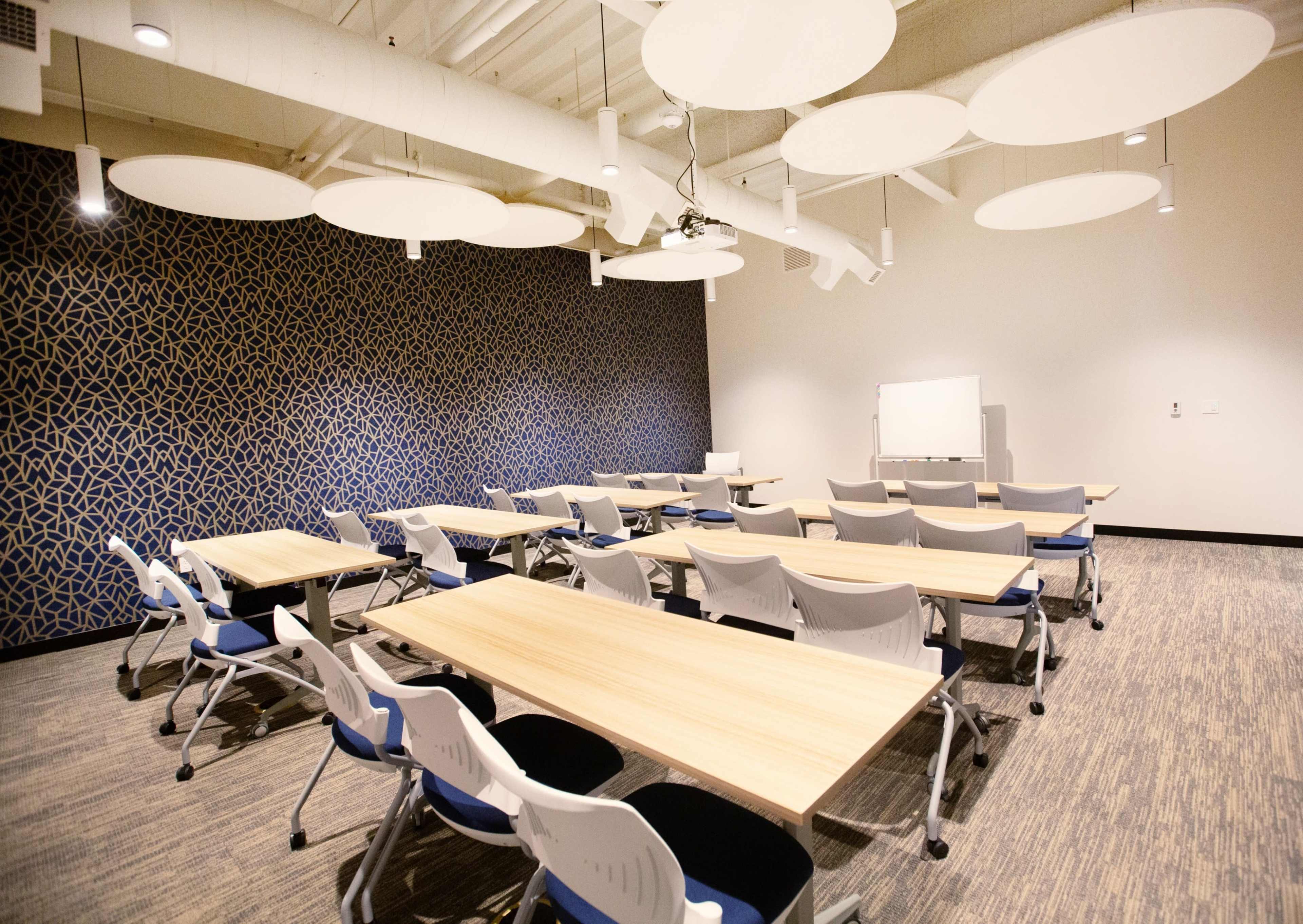 A modern conference room with several tables and chairs arranged for a meeting, featuring a patterned wall and overhead circular light fixtures.