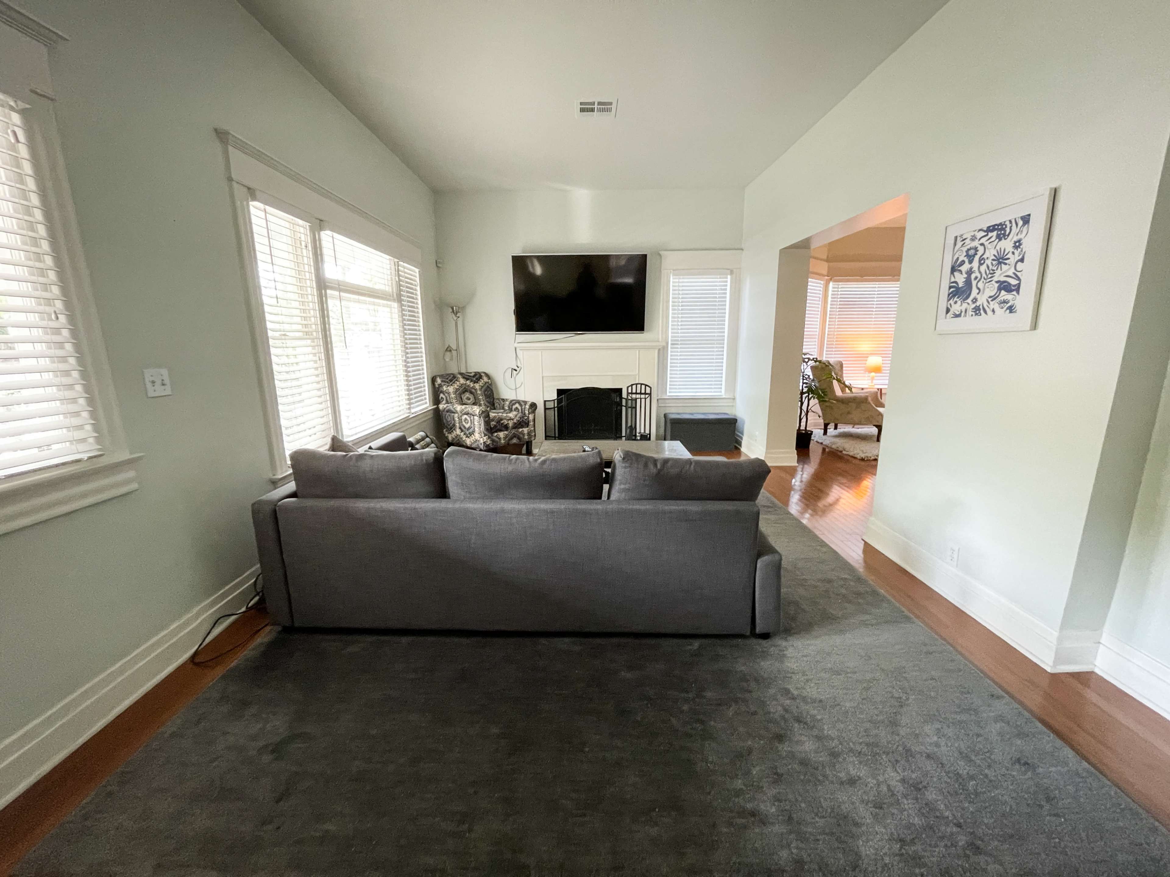 The image shows a living room with a gray sofa facing a wall-mounted television, situated near windows with white blinds.