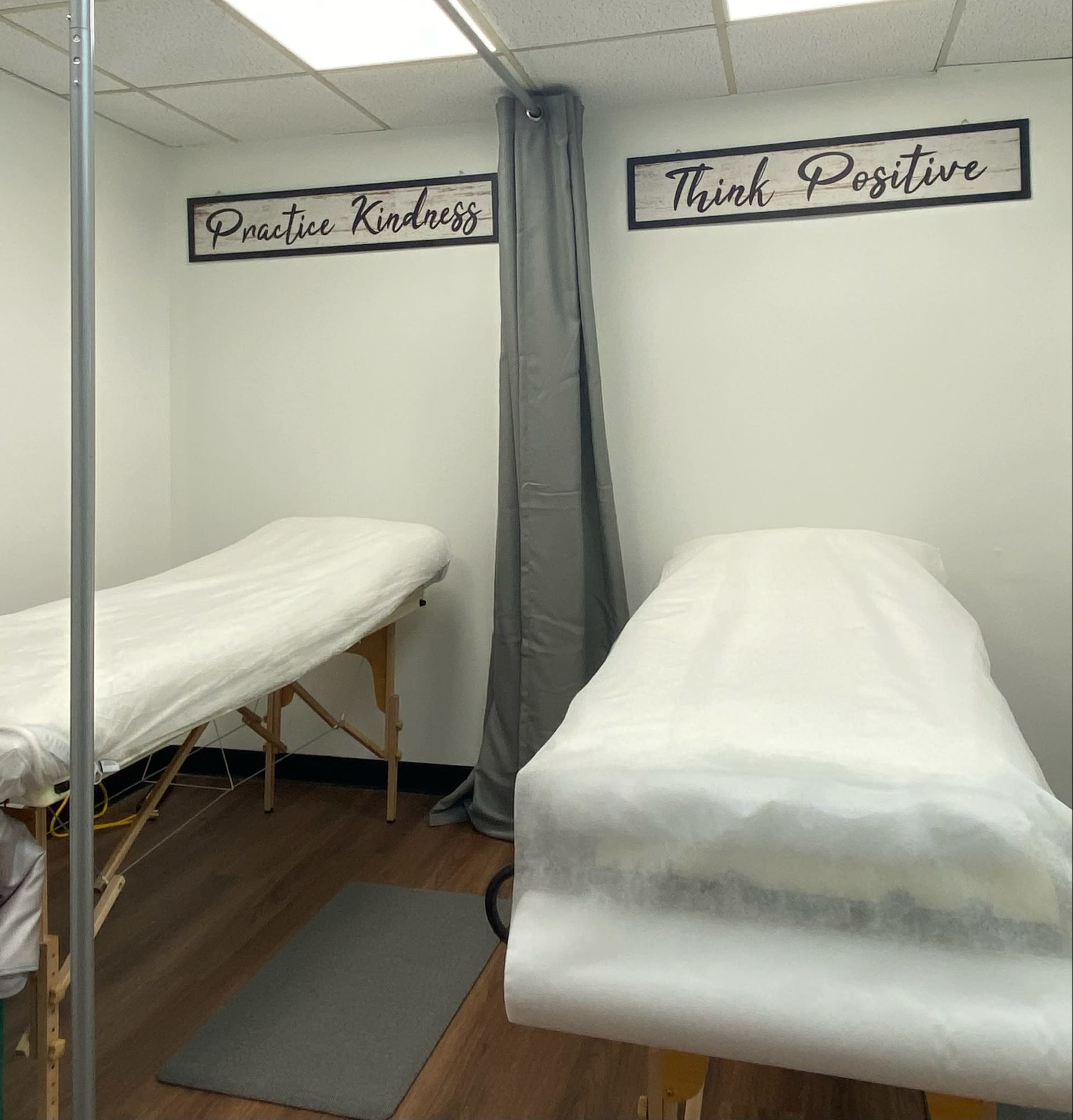 The image shows a massage therapy room with two treatment tables covered in white linens, and motivational signs on the wall that read "Practice Kindness" and "Think Positive."