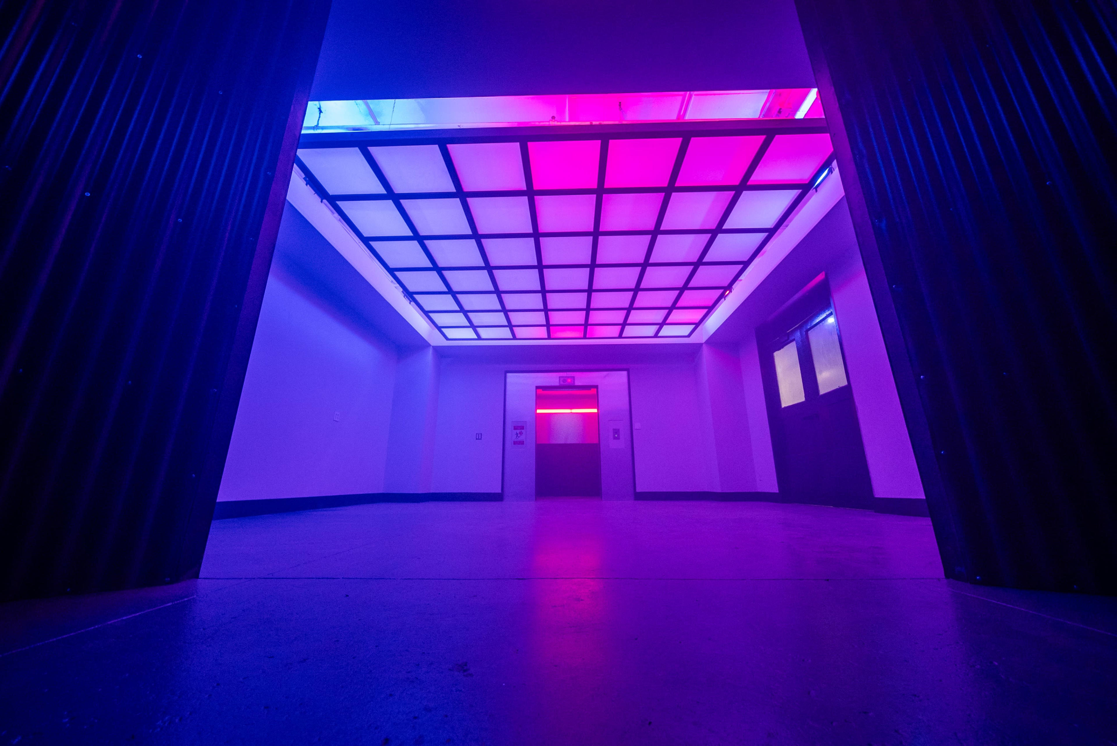 A brightly lit hallway with a futuristic ceiling featuring pink and blue lights, leading to an elevator.