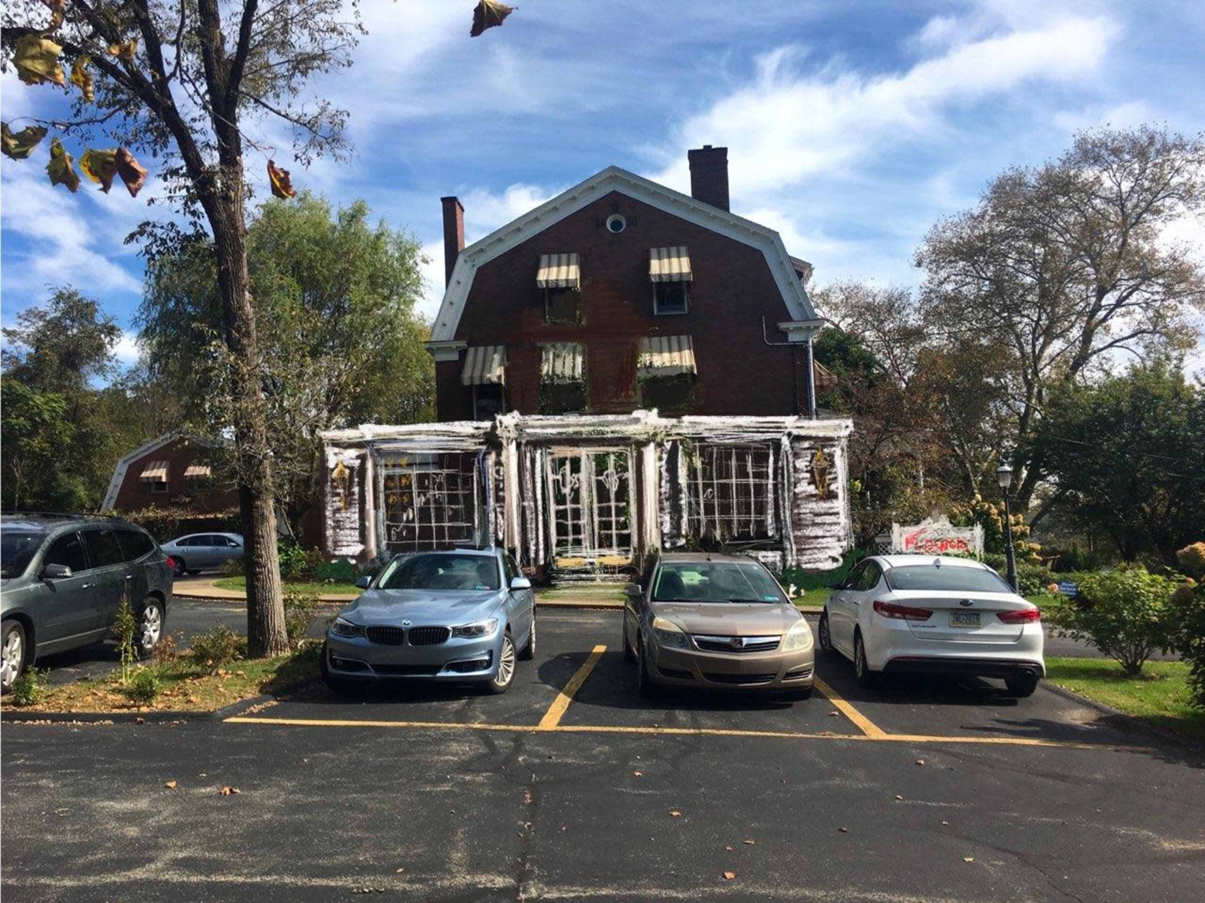 A large brick house with white trim and a porch is situated behind parked cars in a parking lot under a blue sky with scattered clouds.