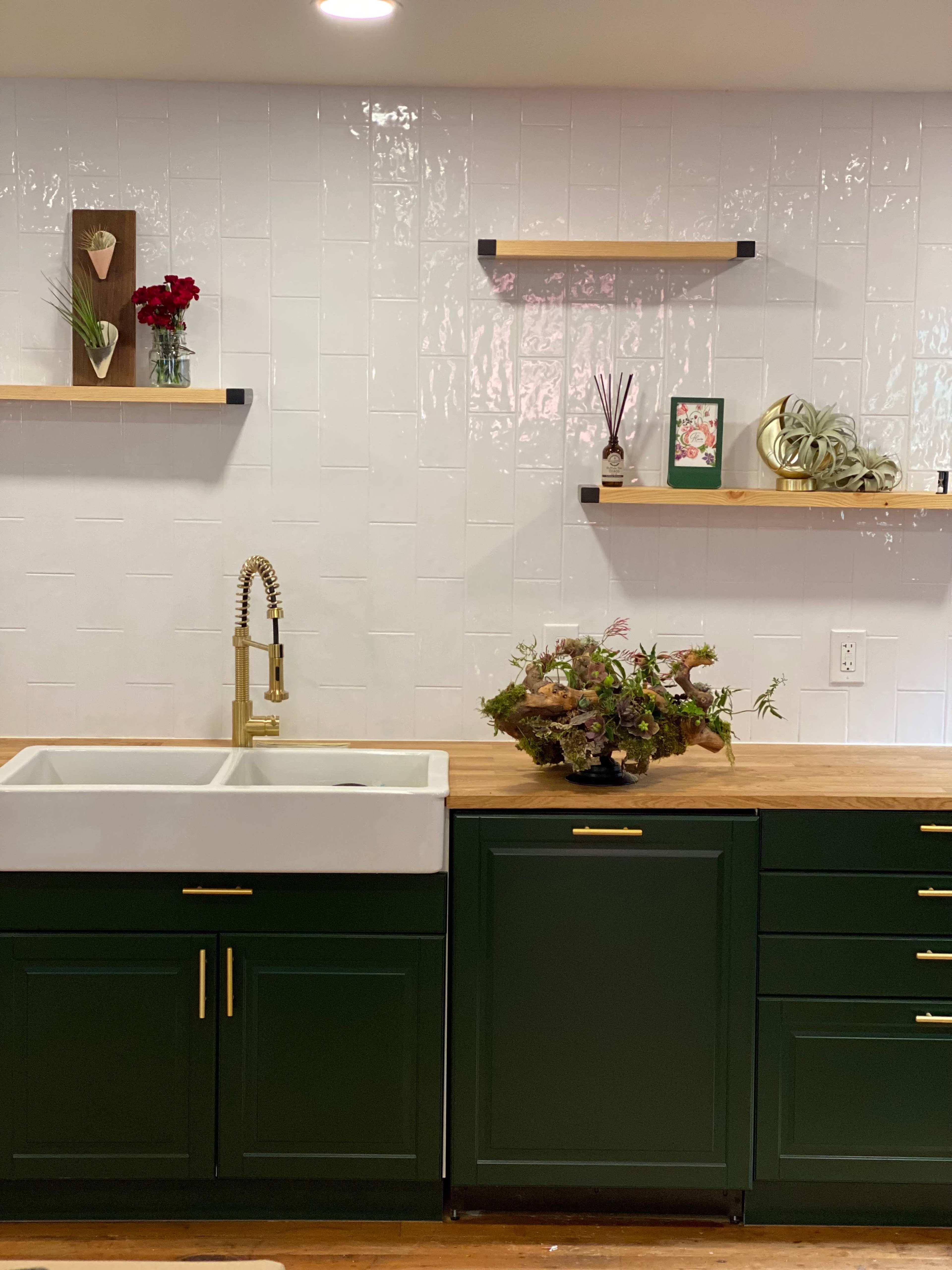 A kitchen with a green base cabinet and a farmhouse sink, accented by wooden shelves displaying decorative items and plants on a white tiled wall.