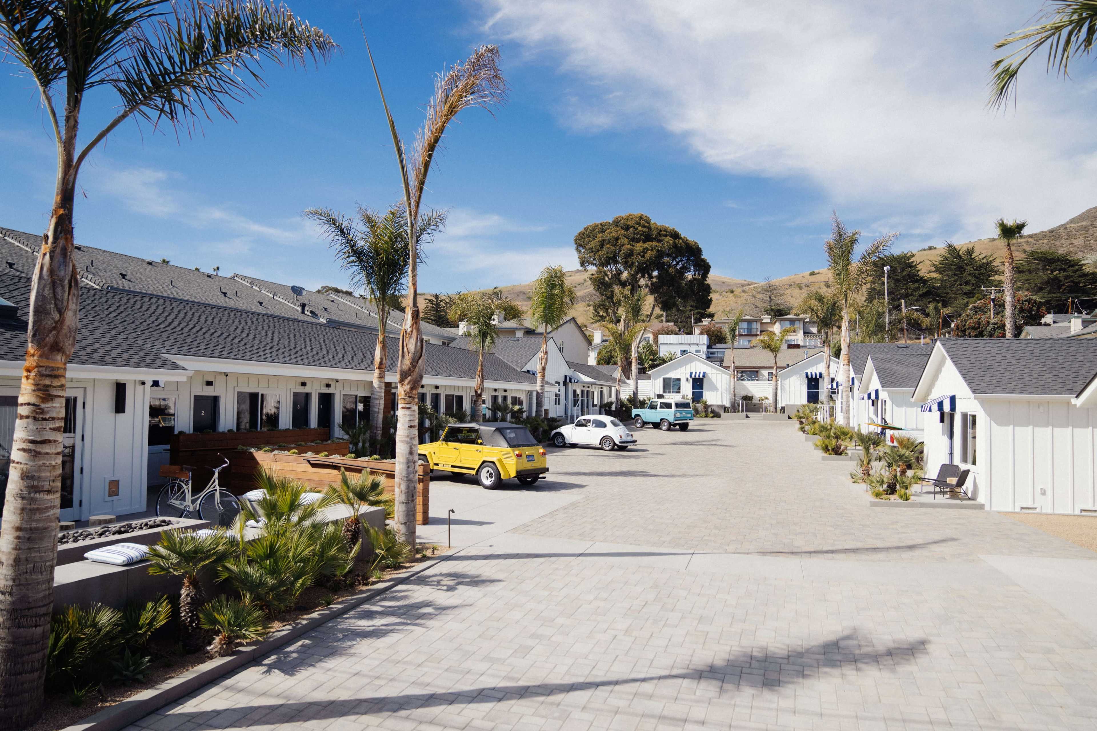 The image shows a renovated courtyard with several white cottages lined on either side, palm trees, and a range of parked cars.