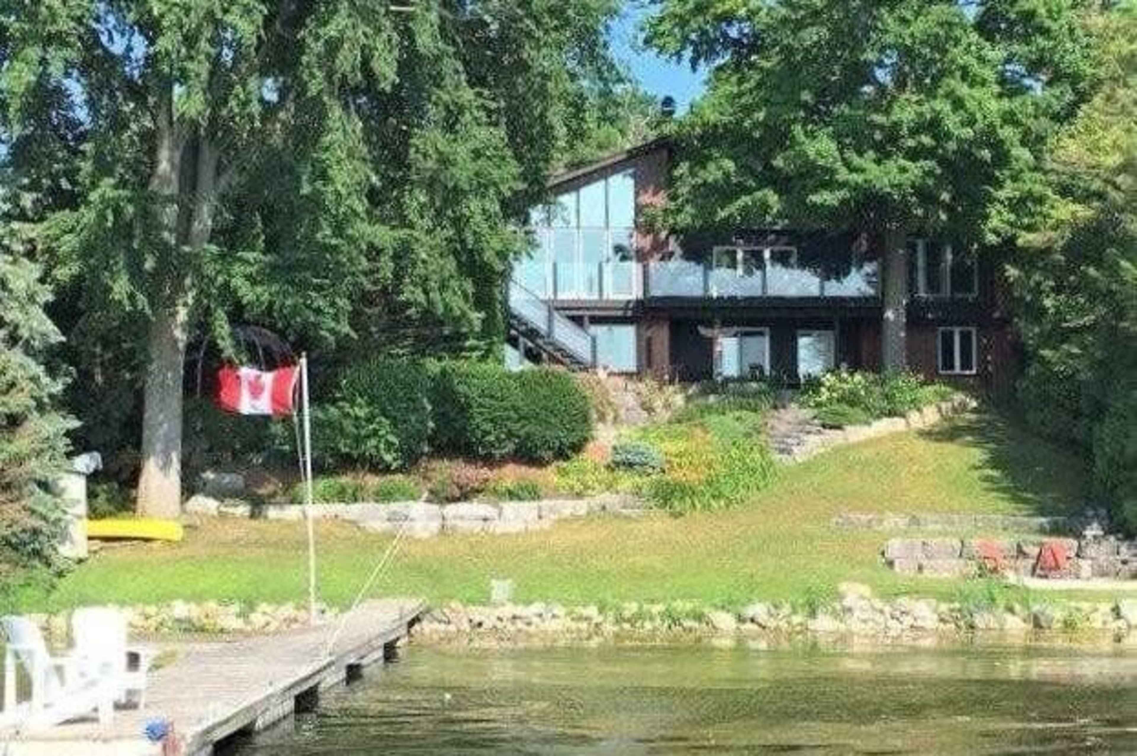 A large house with multiple balconies overlooks a lake, surrounded by greenery and a Canadian flag near a wooden dock.