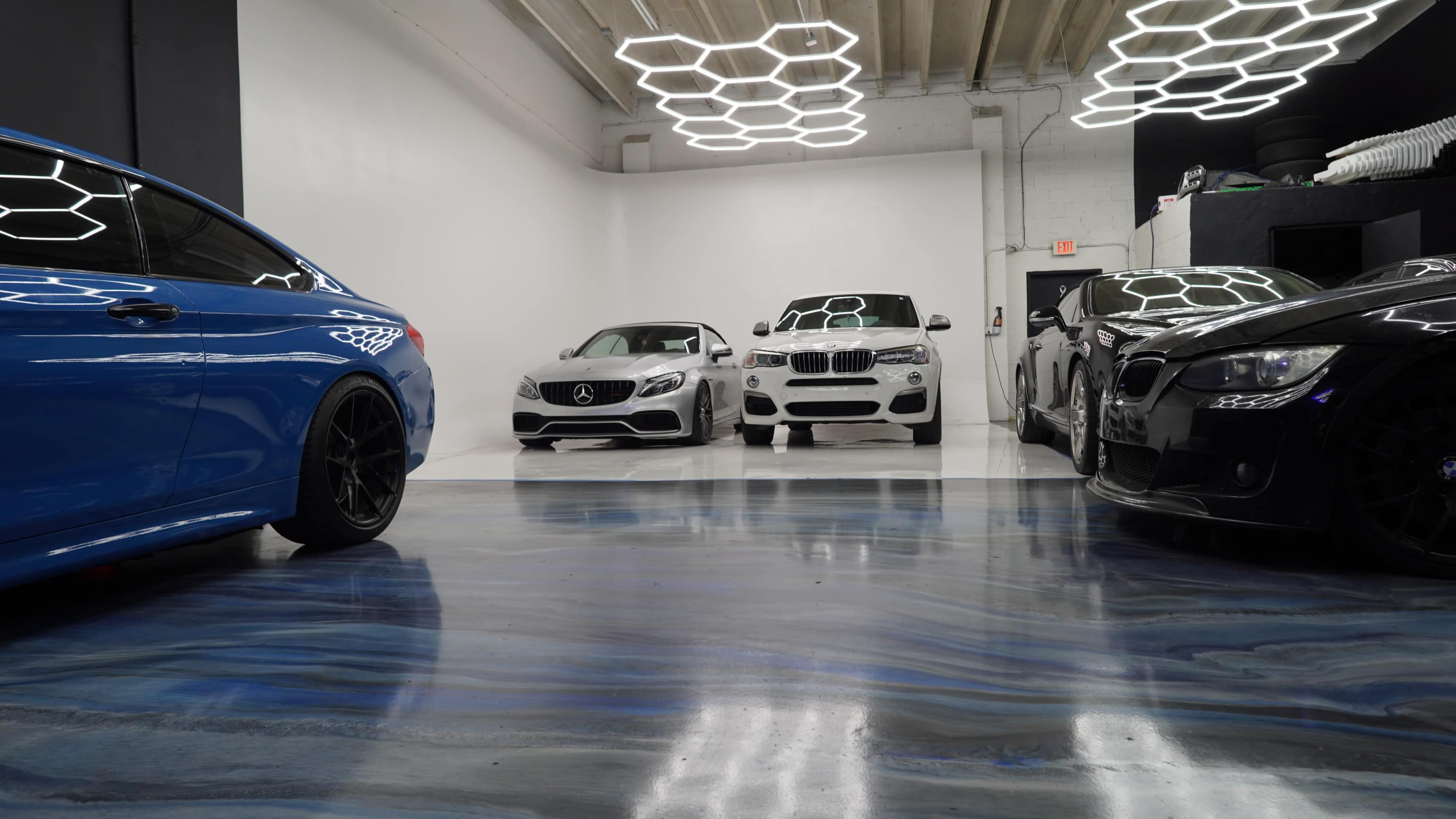 The image shows a modern car showroom featuring a blue car on the left, a silver Mercedes-Benz in the center, a white BMW next to it, and a black car on the right, all on a polished blue-and-gray floor.
