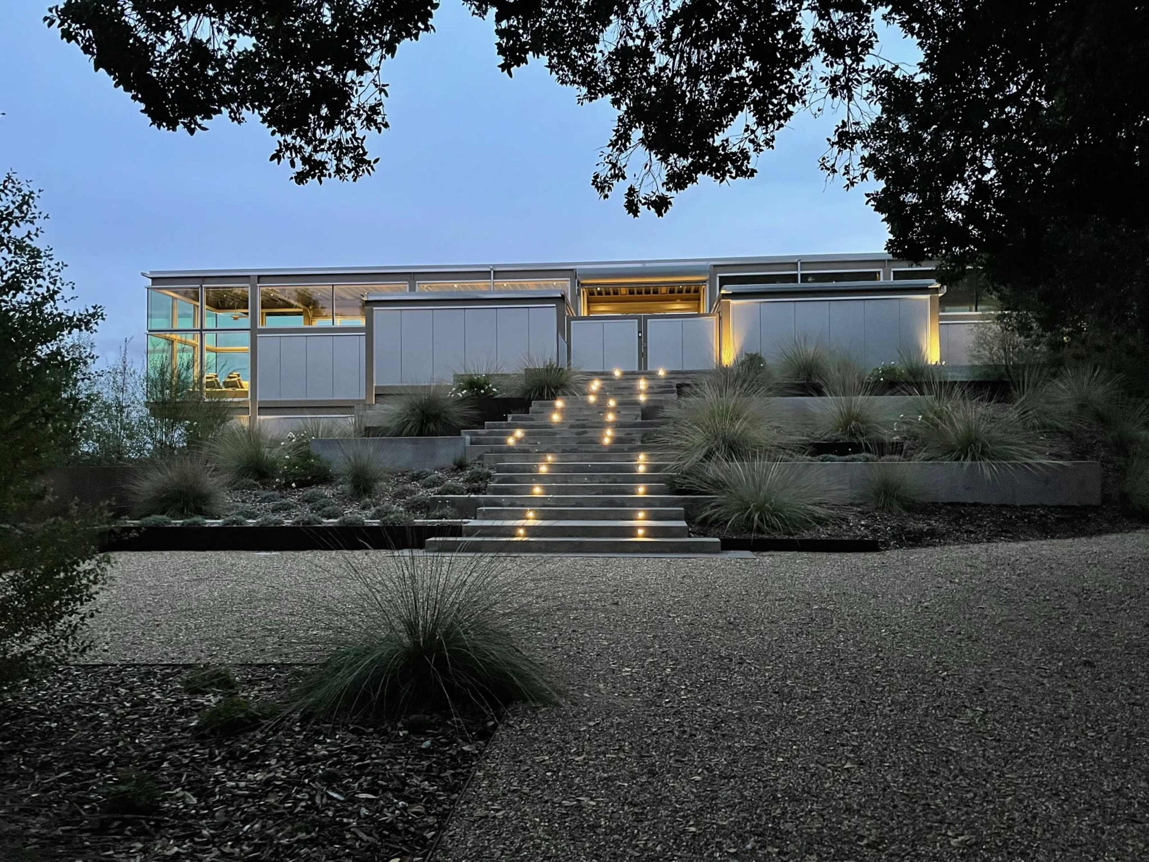 A modern house is illuminated by soft outdoor lighting near a stepped pathway leading up from a gravel driveway, surrounded by landscaped vegetation.