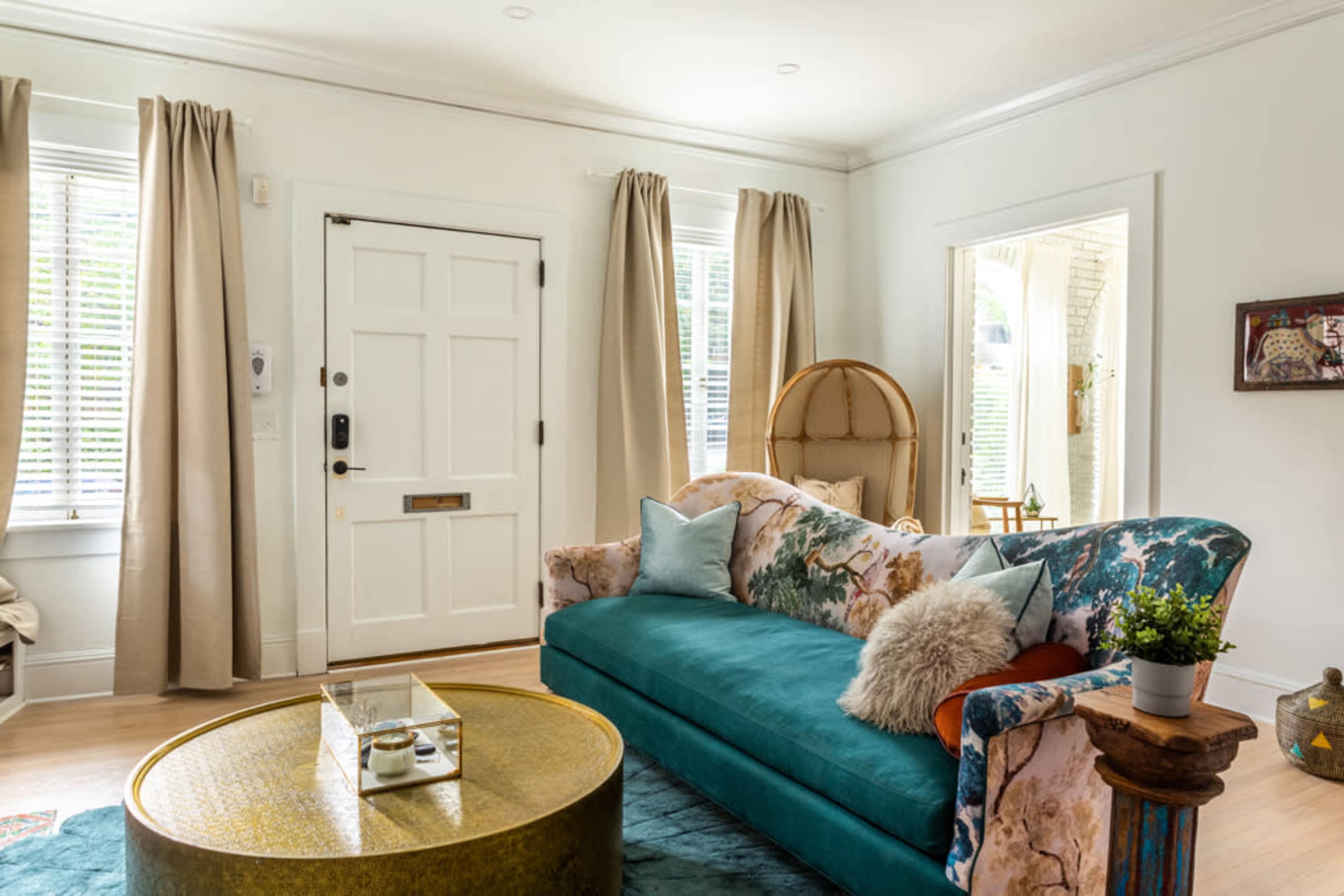 The image shows a living room featuring a blue velvet sofa with floral patterns, a round gold coffee table, and natural light coming through large windows with beige curtains.