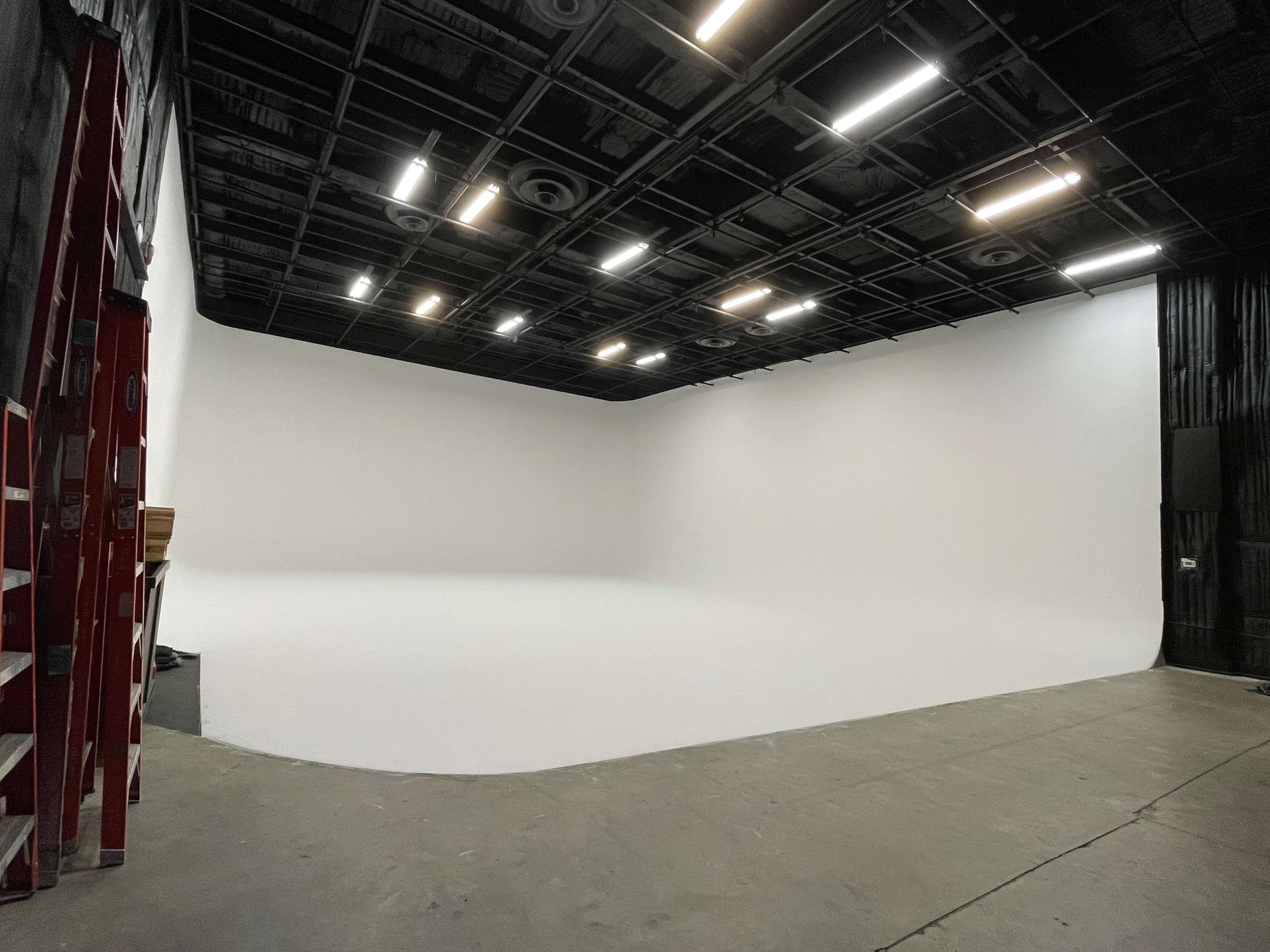 The image shows a large, empty studio space with a curved white backdrop and overhead lighting.