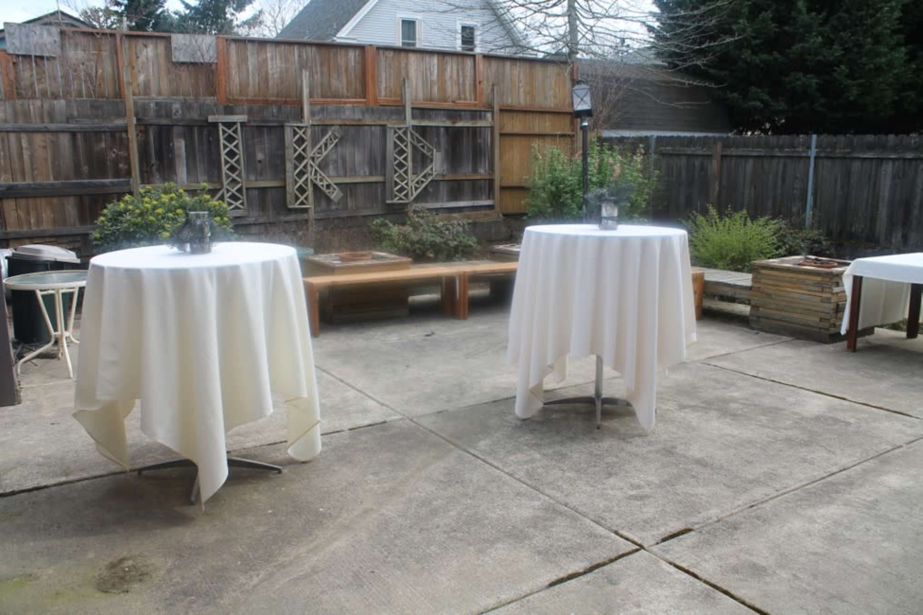 A patio with two round tables covered in white tablecloths, surrounded by a wooden fence and greenery.