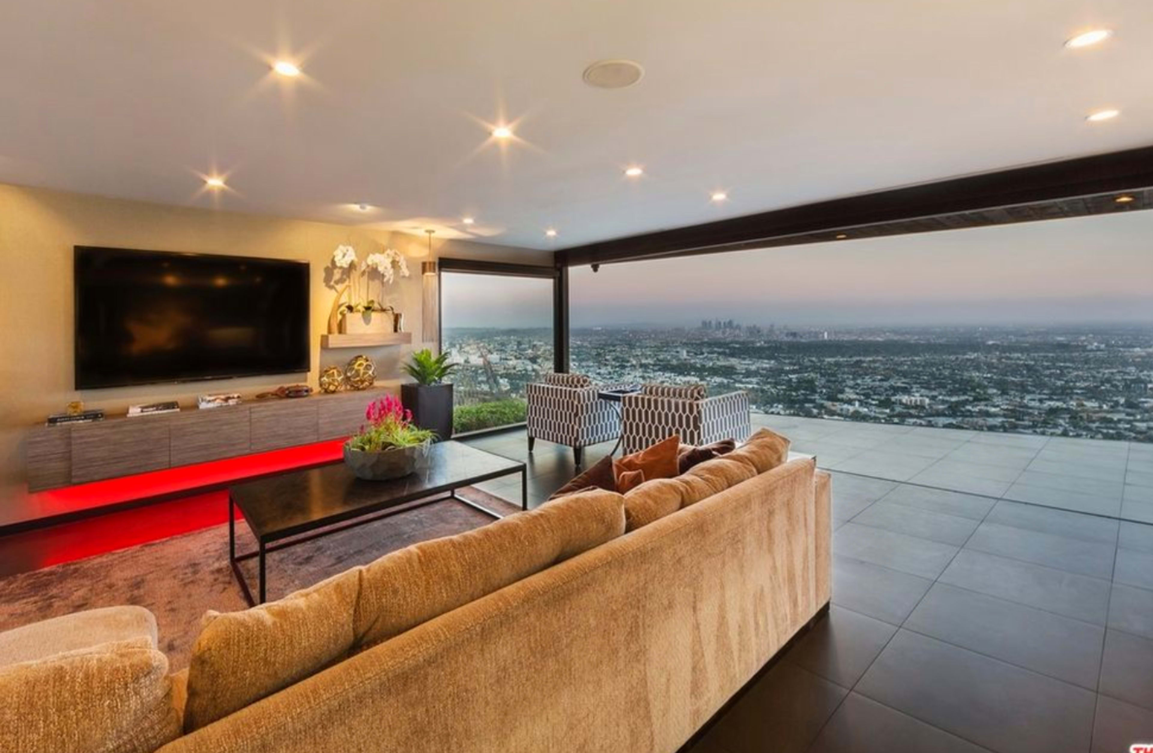 The image shows a modern living room that opens up to a panoramic view of a city skyline through large glass doors.