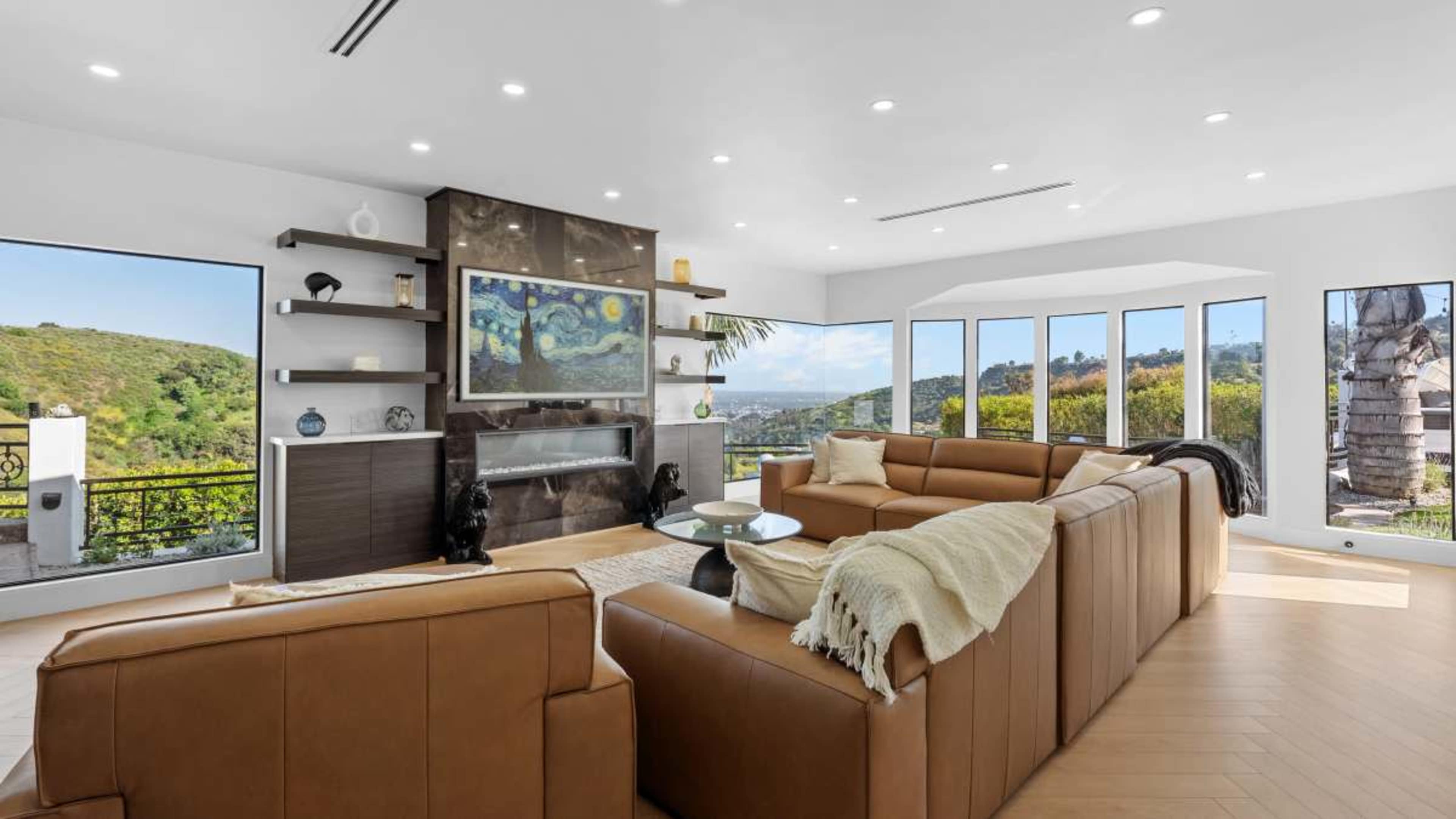 The image features a modern living room with large windows, a sleek fireplace, and brown leather couches arranged around a glass coffee table.