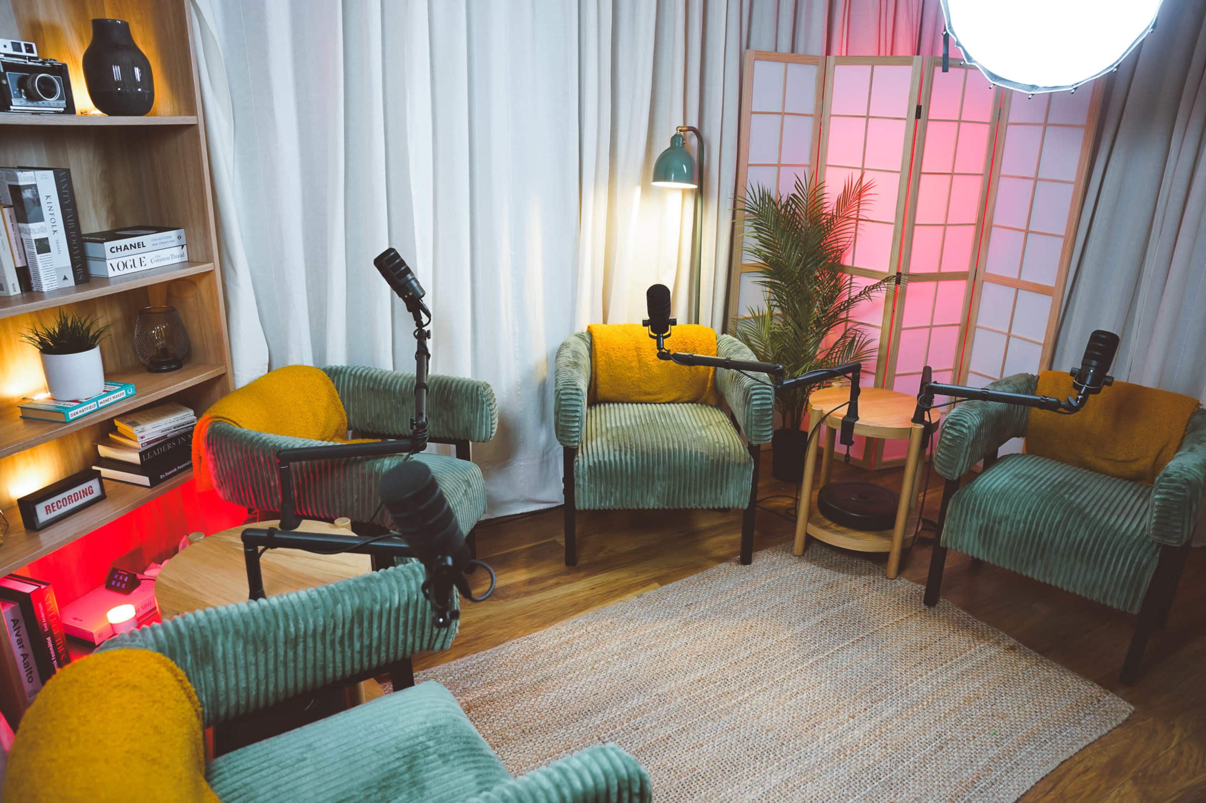 The image shows a cozy podcast studio setup with four chairs, each equipped with a microphone, surrounded by a bookshelf and decorative plants.